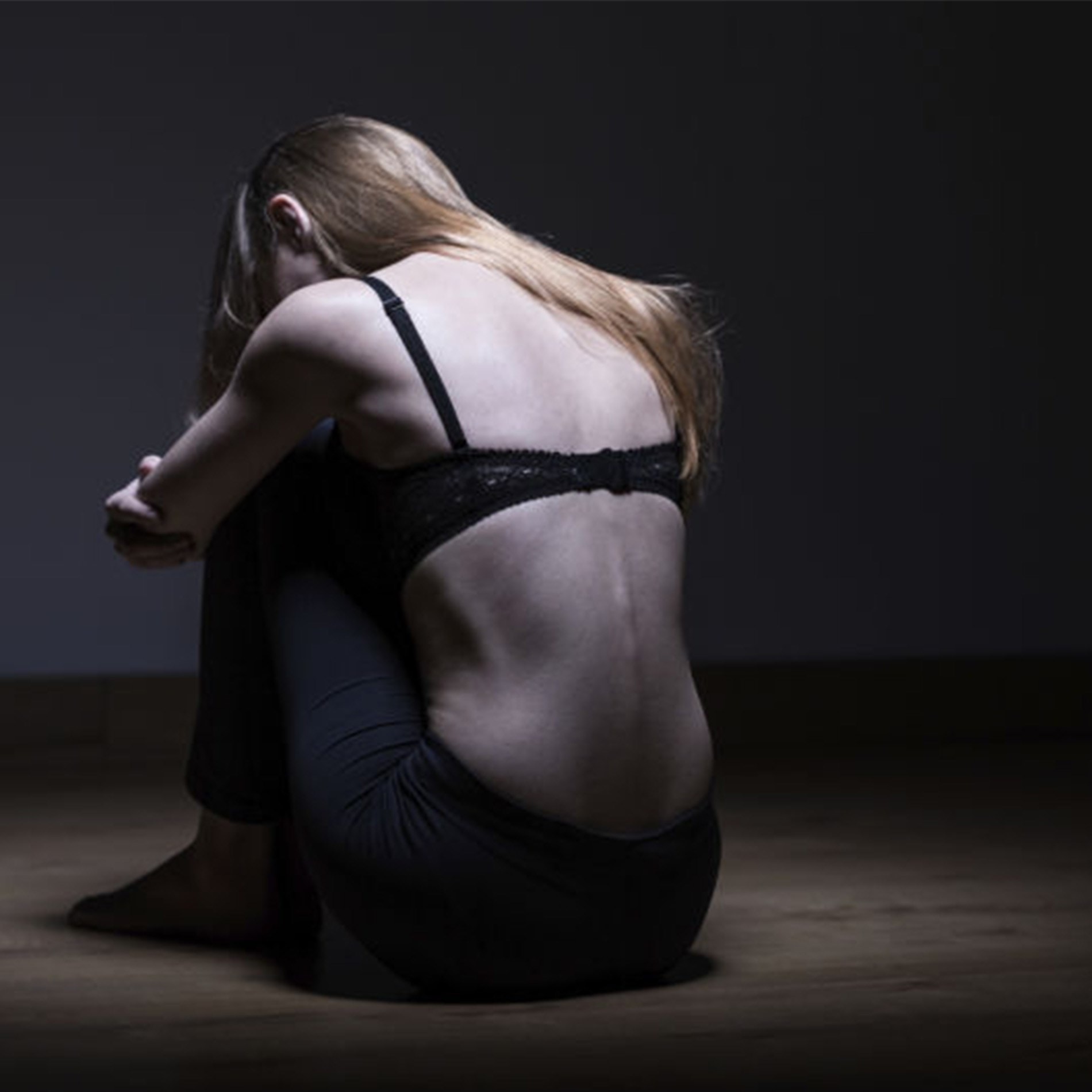 A woman with long hair sits on the floor in a dimly lit room, hugging her knees, wearing a black top and pants, facing away.