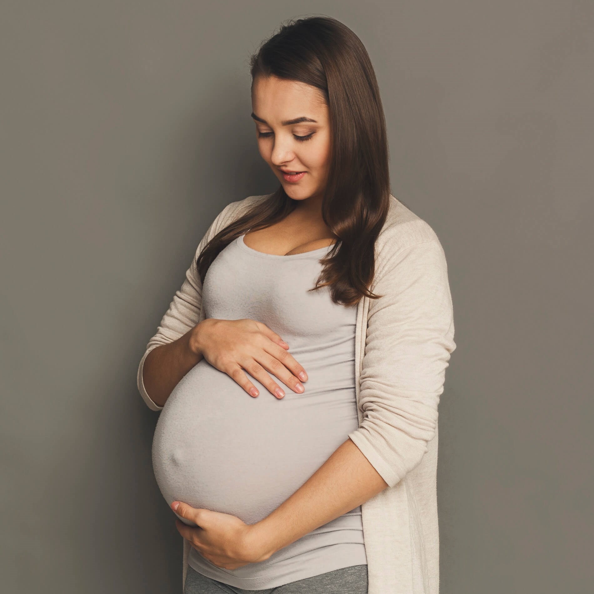 Pregnant woman in a light cardigan and gray top, gently cradling her belly, stands against a gray background, looking down with a soft smile.