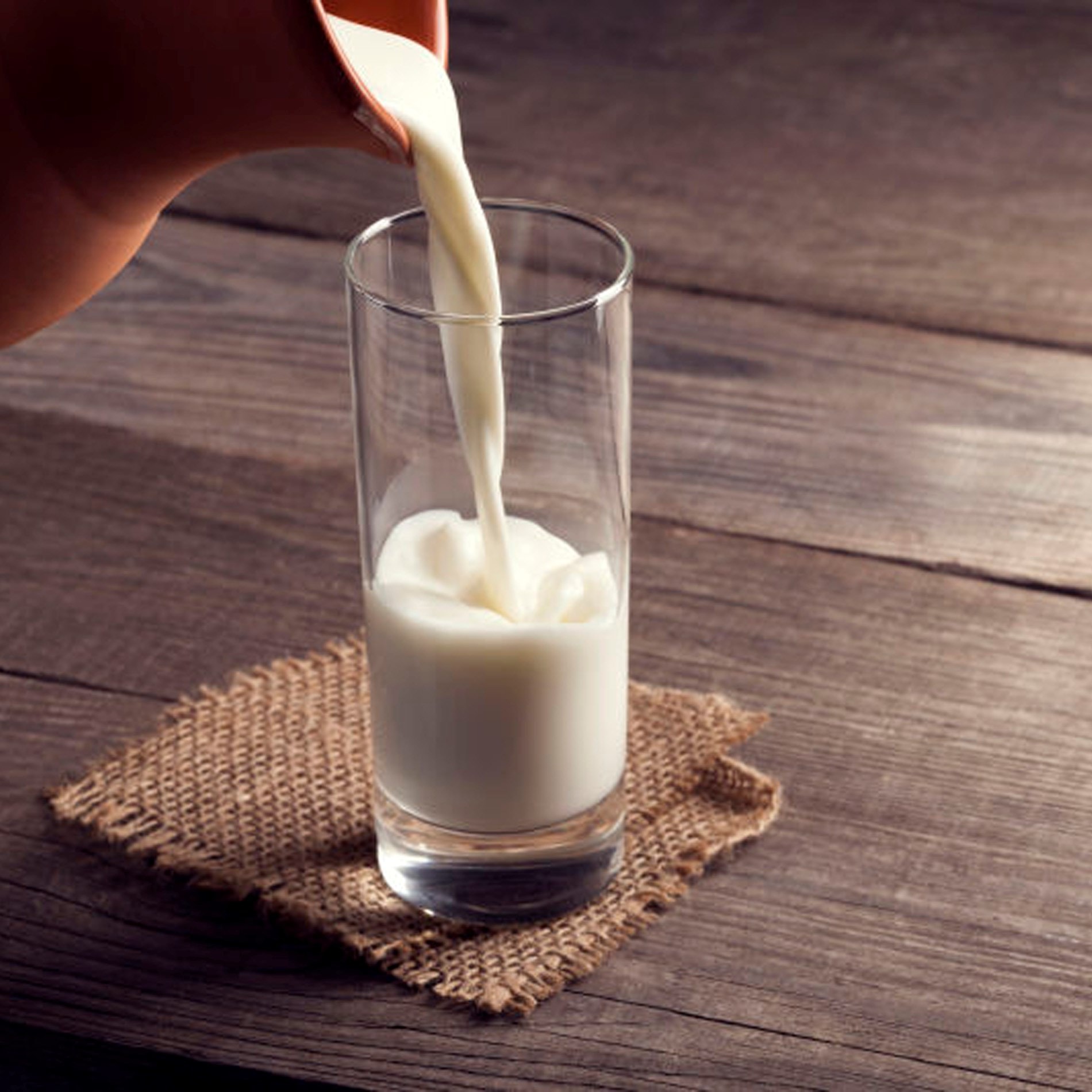 Milk being poured into a clear glass on a burlap mat, set on a rustic wooden table.