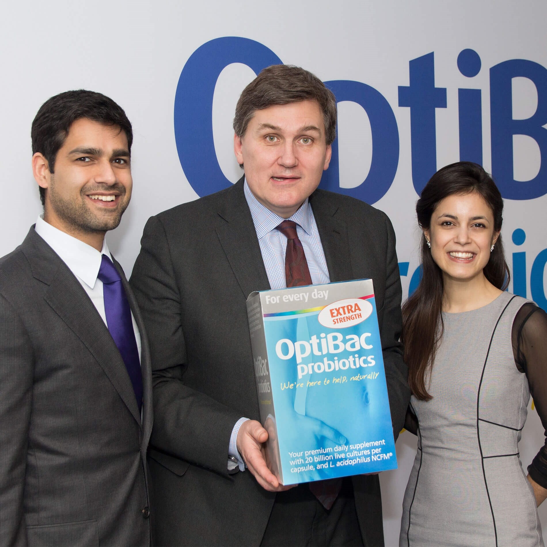 Three people smiling, one holding a large OptiBac probiotics box. They stand in front of a backdrop with the OptiBac logo.