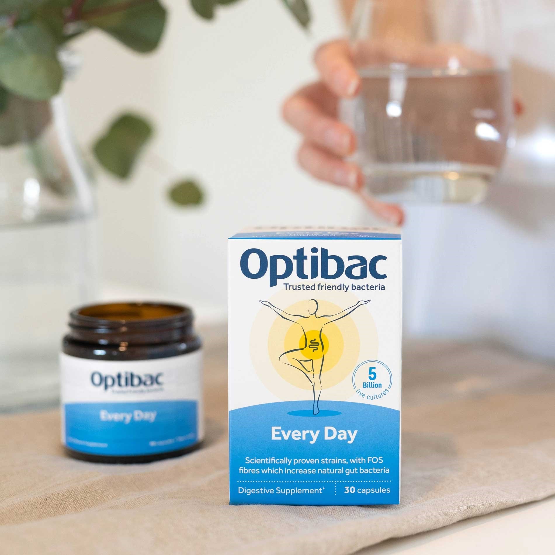 Optibac Every Day probiotic box and jar on a table, with a hand holding a glass of water in the background.