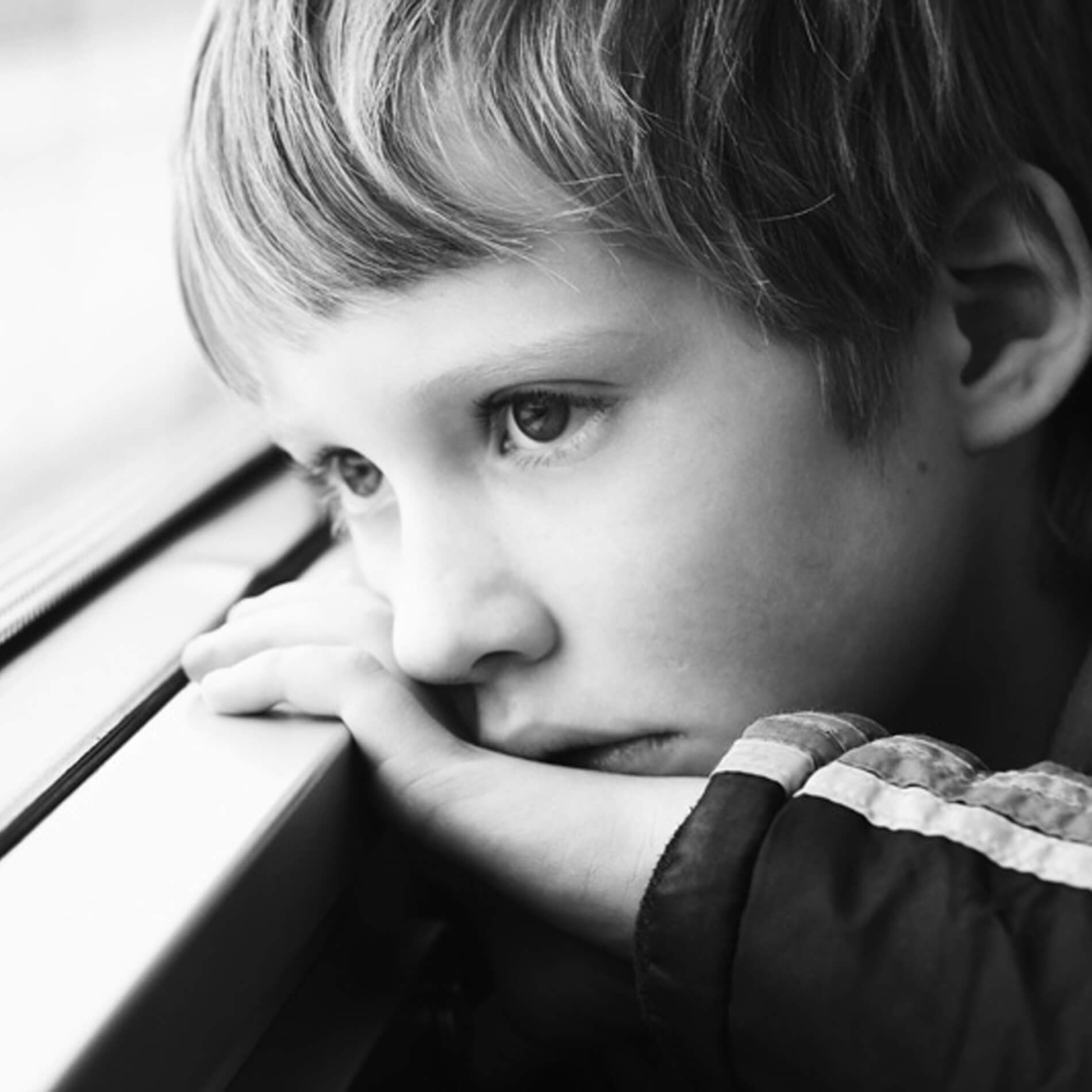 Black and white image of a young boy gazing out a window, resting his chin on his hand, with a thoughtful expression.