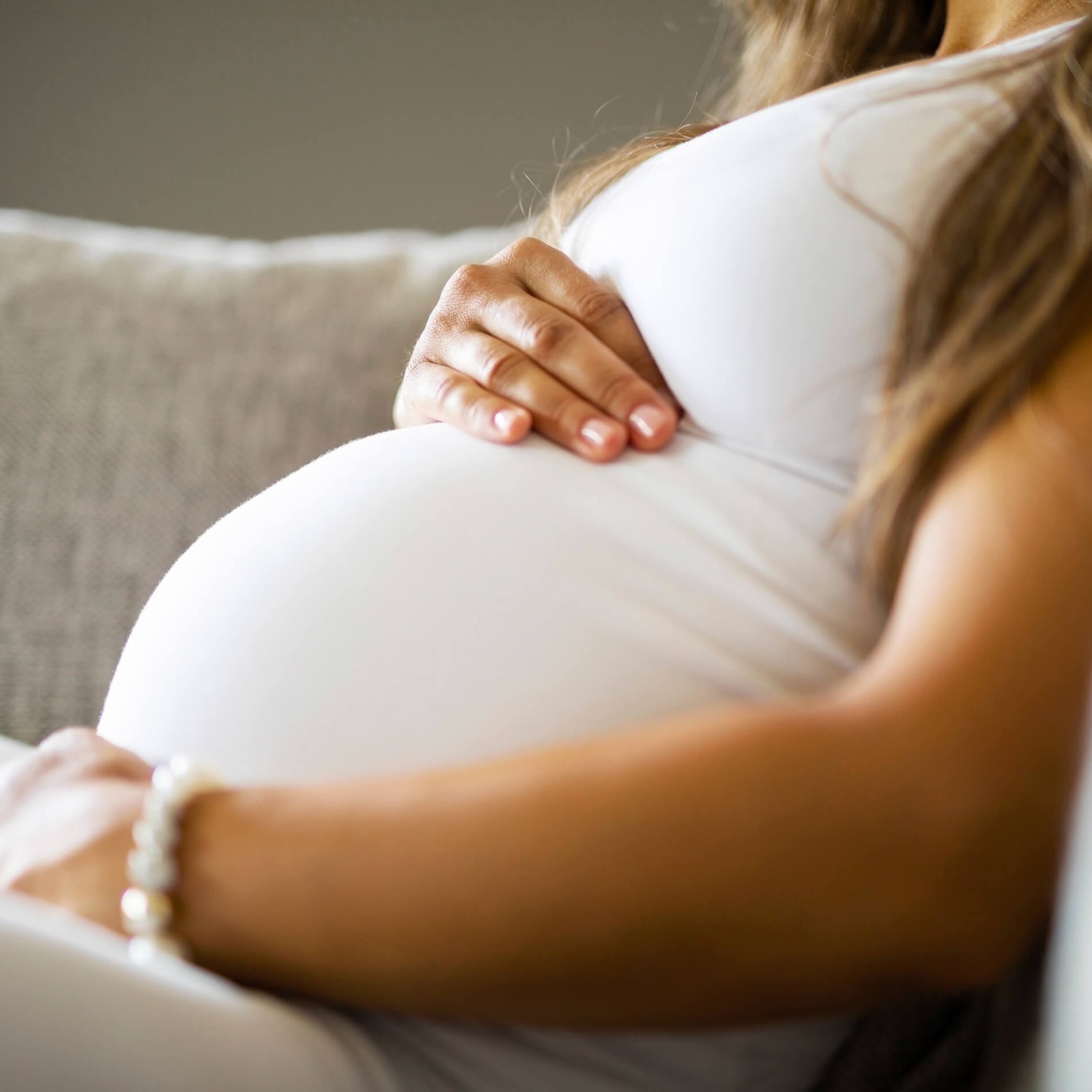 Pregnant woman in a white dress sitting on a couch, gently cradling her belly with one hand, wearing a bracelet on her wrist.