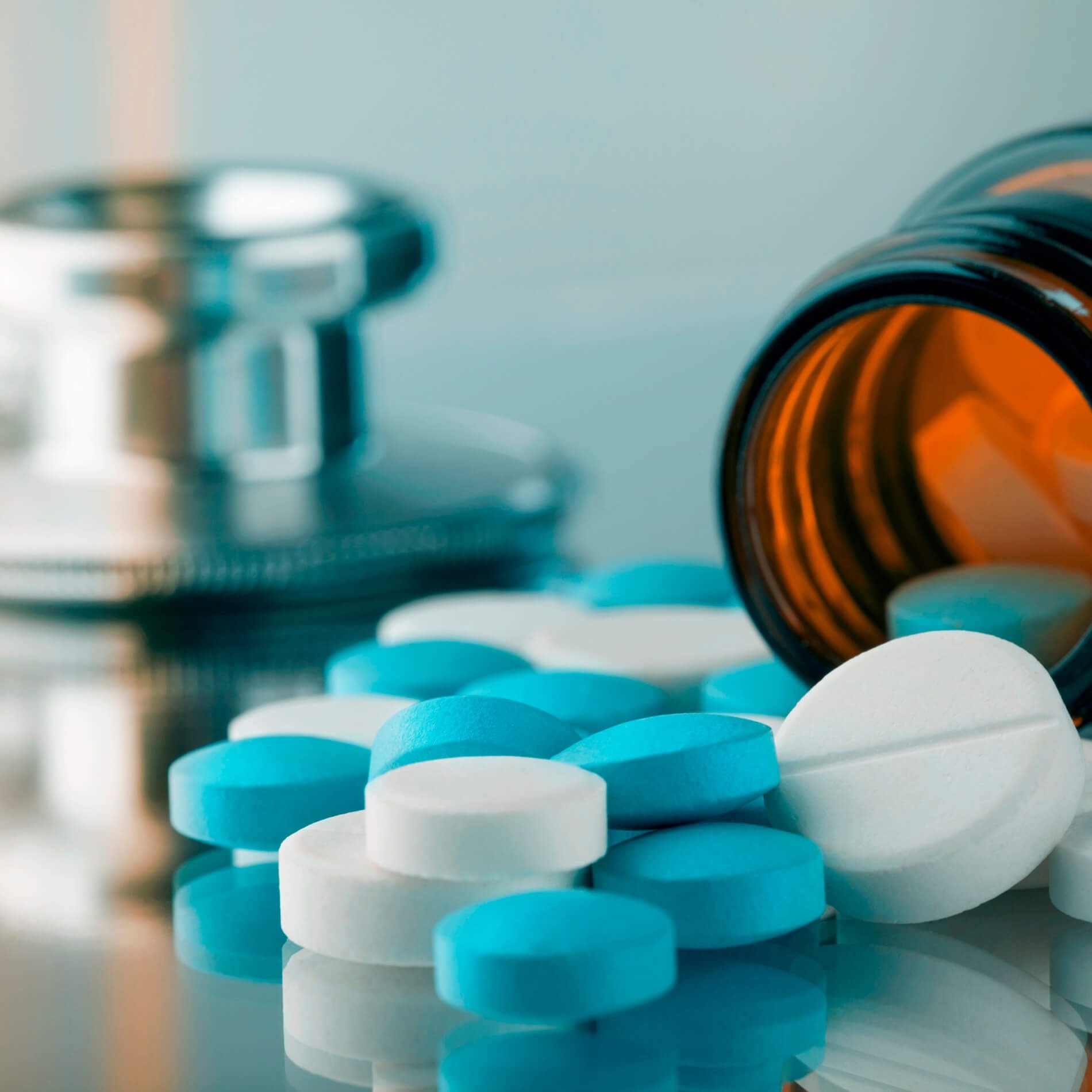Close-up of blue and white pills scattered from a brown bottle next to a stethoscope on a reflective surface.