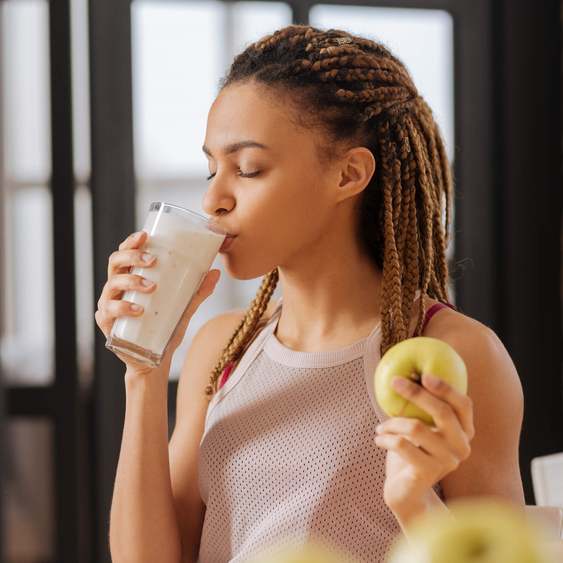Woman with braided hair drinks a smoothie while holding a green apple, wearing a sleeveless top in a bright room.