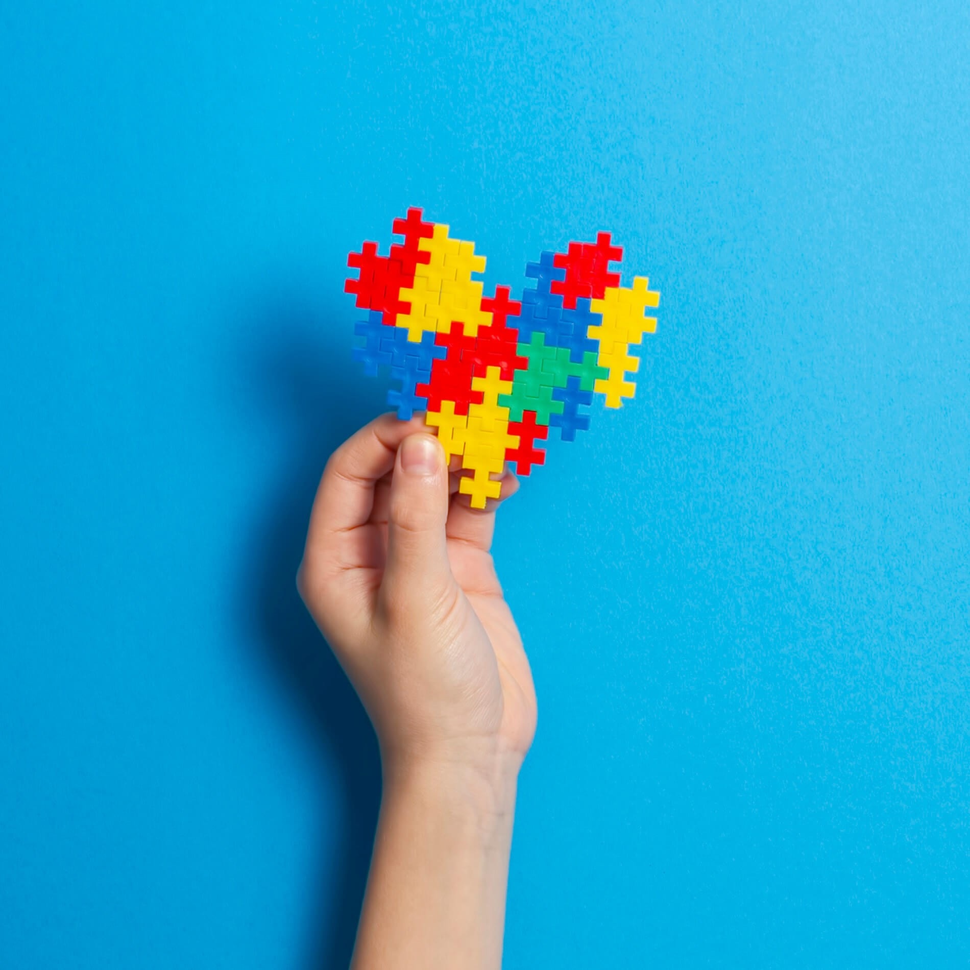 A hand holds a heart-shaped puzzle made of colorful interlocking pieces against a bright blue background.