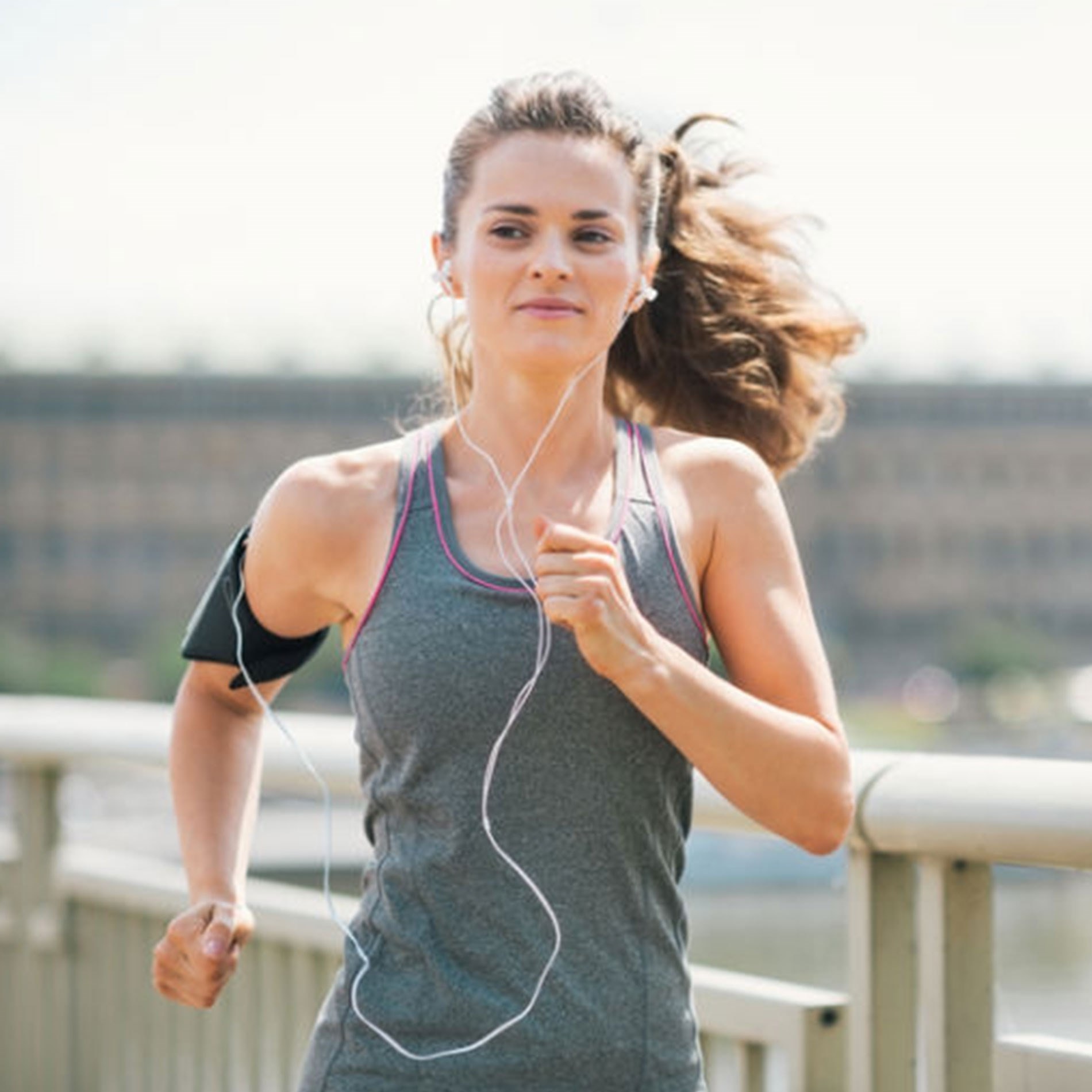Woman jogging outdoors, wearing a gray tank top and earphones, with an armband. She has her hair tied back and is smiling.