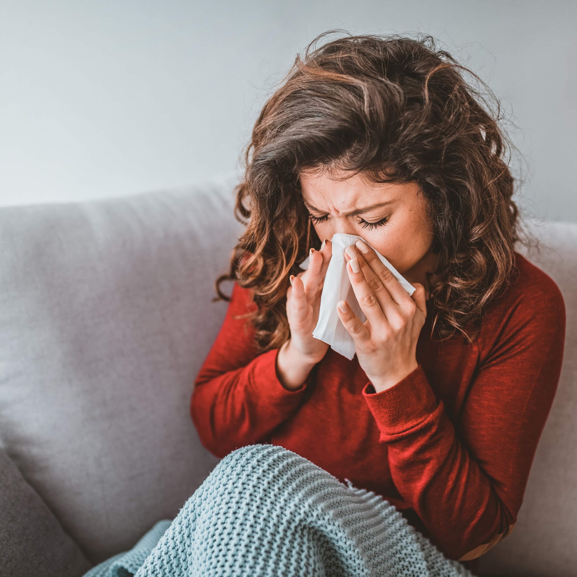 Woman with curly hair sitting on a couch, sneezing into a tissue, wearing a red sweater and wrapped in a blue blanket.