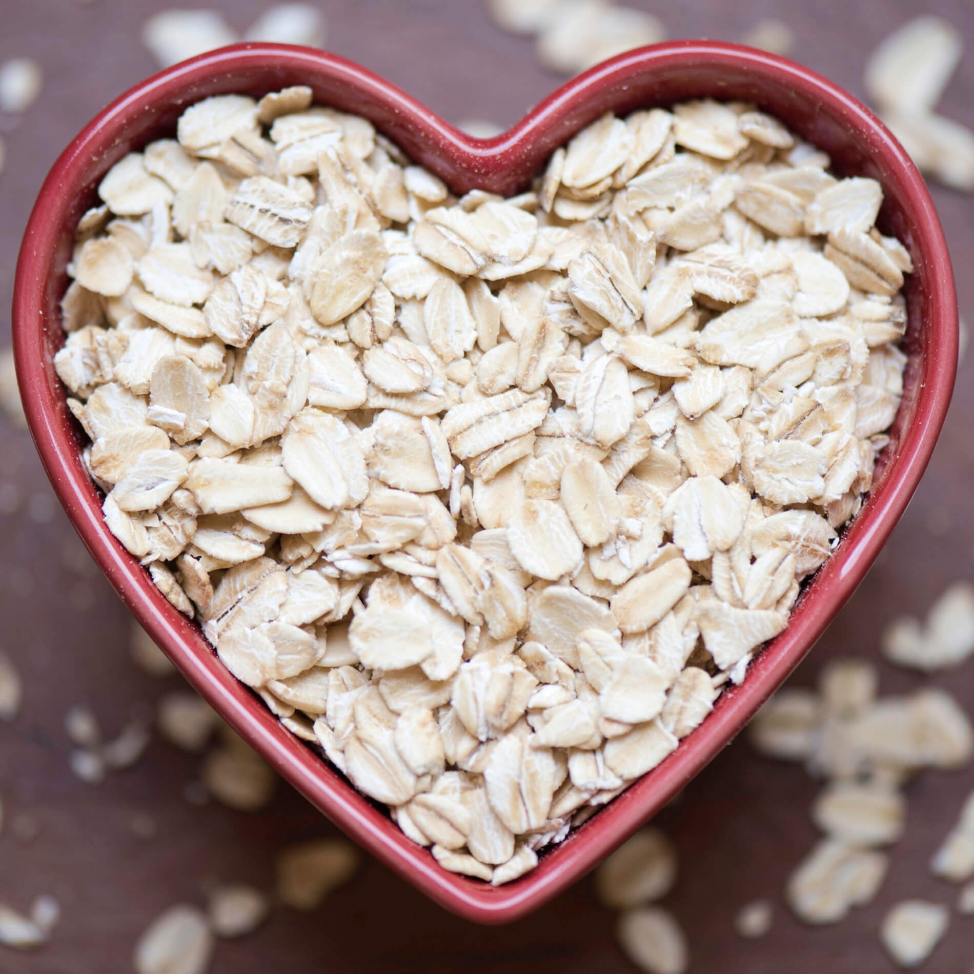 Heart-shaped bowl filled with rolled oats, placed on a brown surface with scattered oats around.