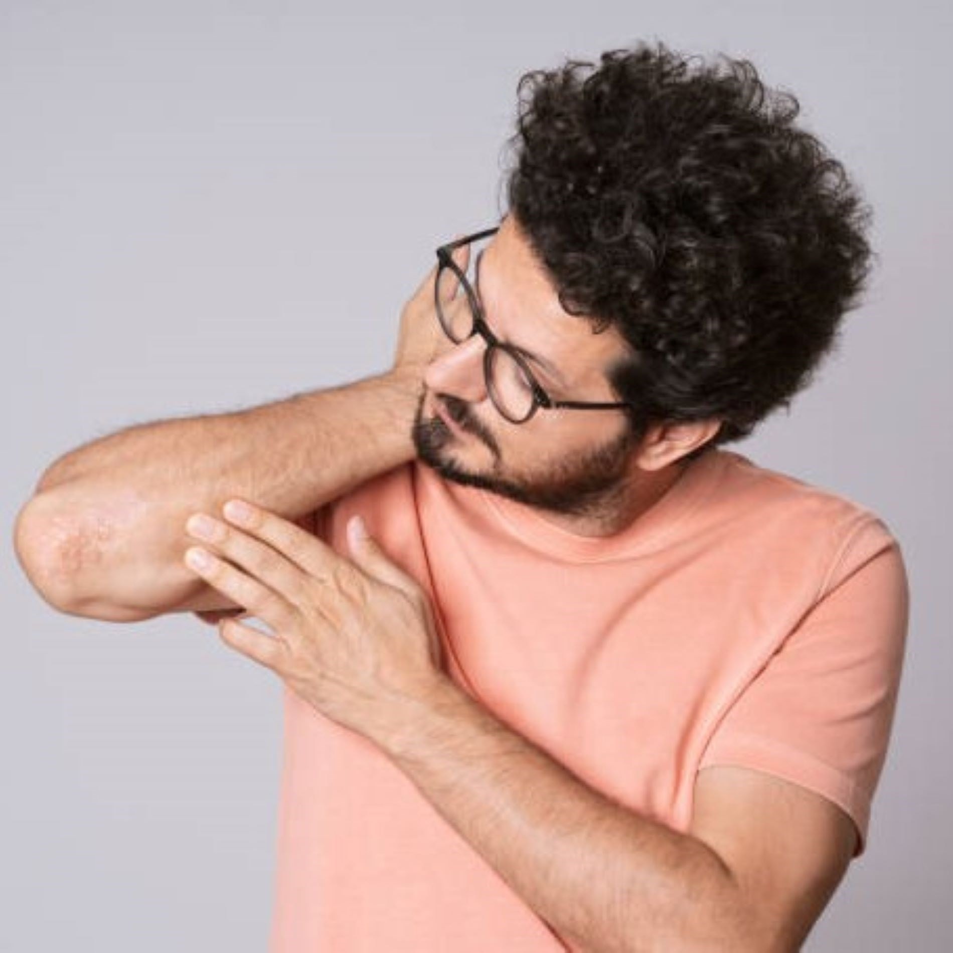 Man in a peach shirt and glasses examines his irritated elbow, showing visible skin redness, against a gray background.