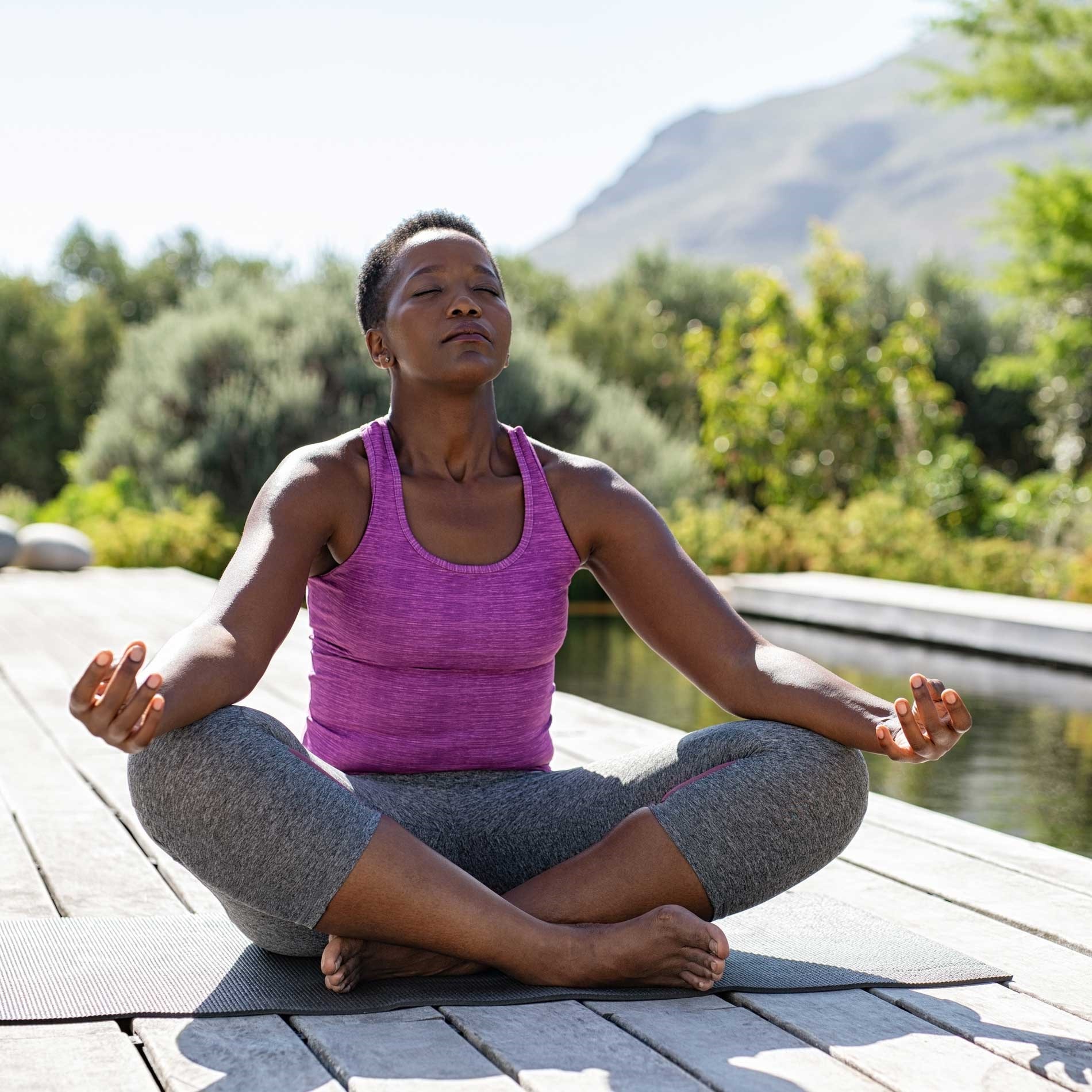 A person sits cross-legged on a yoga mat outdoors, meditating with closed eyes, wearing a purple tank top and gray pants.