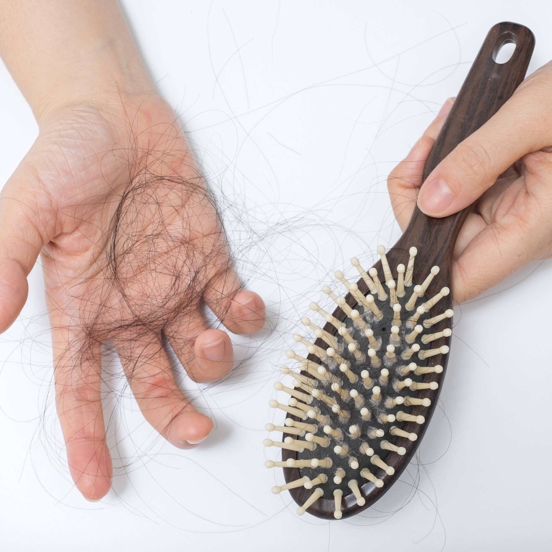 A hand holding a wooden hairbrush filled with hair, and another hand showing a clump of hair against a white background.