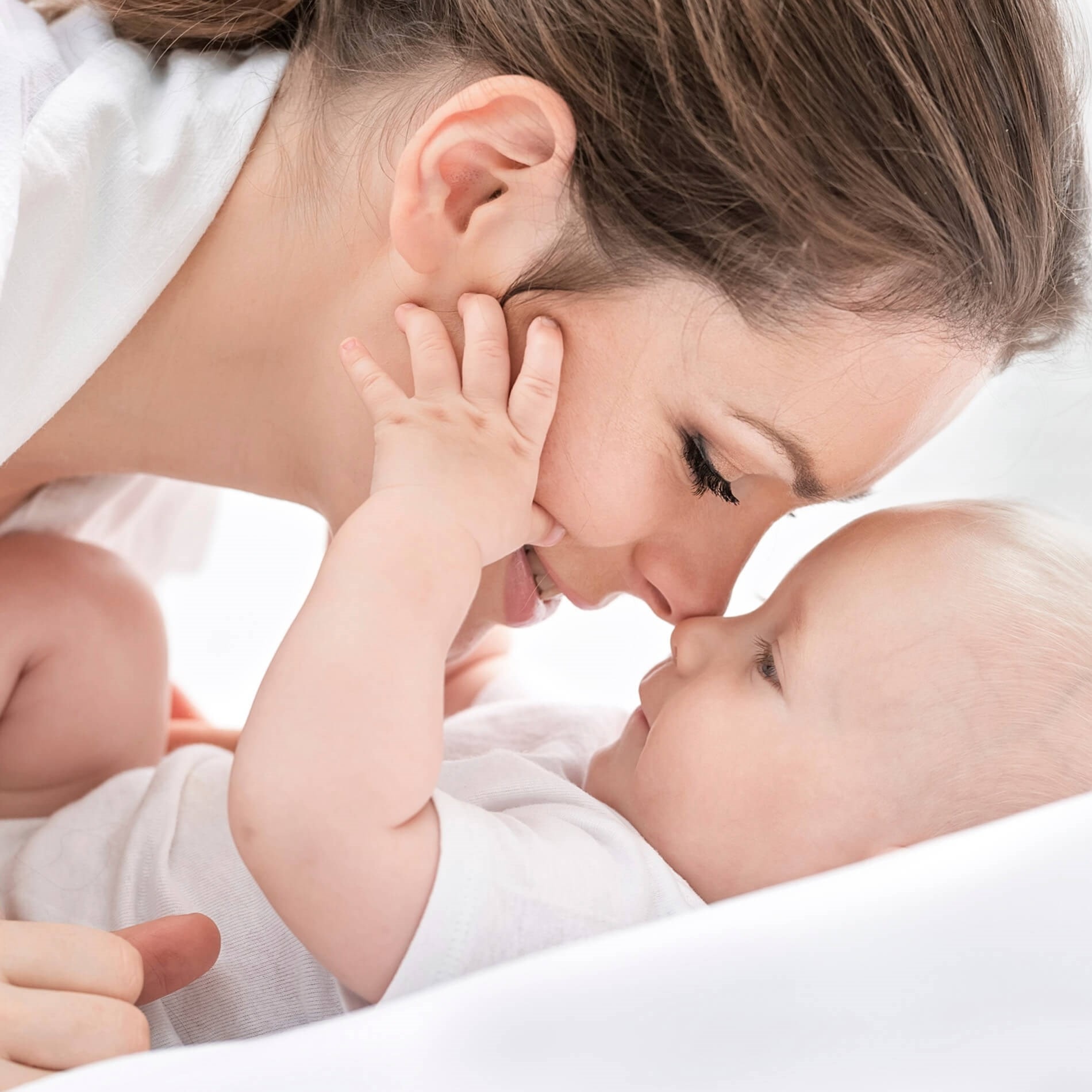 Mother and baby lying face-to-face, smiling and touching noses, both wearing white clothing in a soft, bright setting.