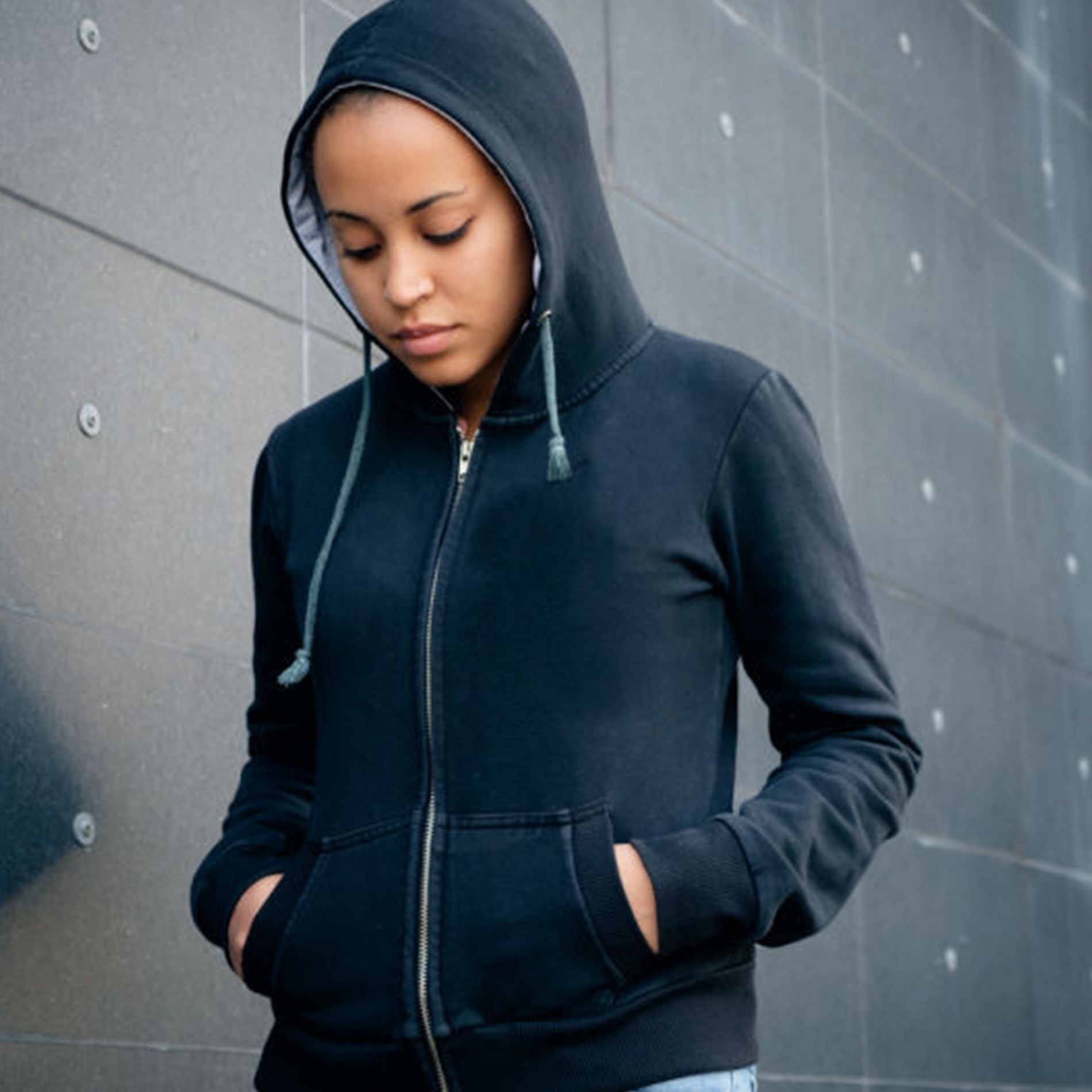 Person in a black hoodie stands against a dark, tiled wall, looking down with hands in pockets.
