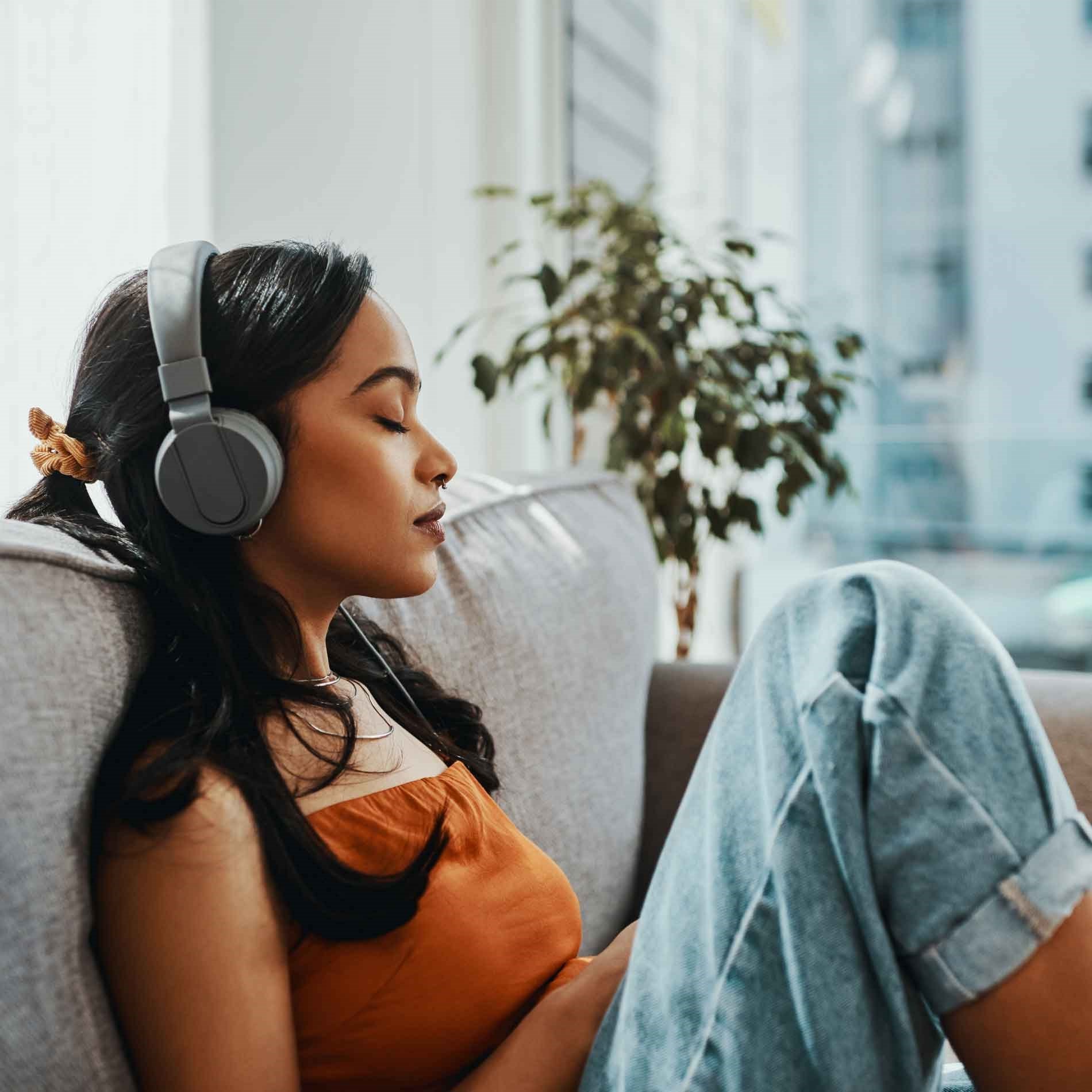 Woman in an orange top and jeans, sitting on a couch with eyes closed, wearing headphones, next to a window with a plant in the background.