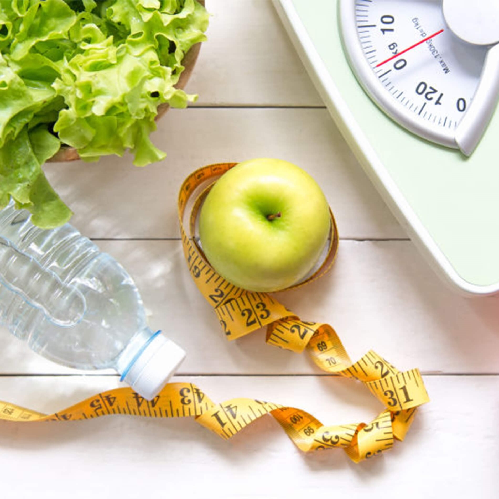 A green apple, lettuce, water bottle, measuring tape, and scale on a white wooden surface, symbolizing healthy living and weight management.