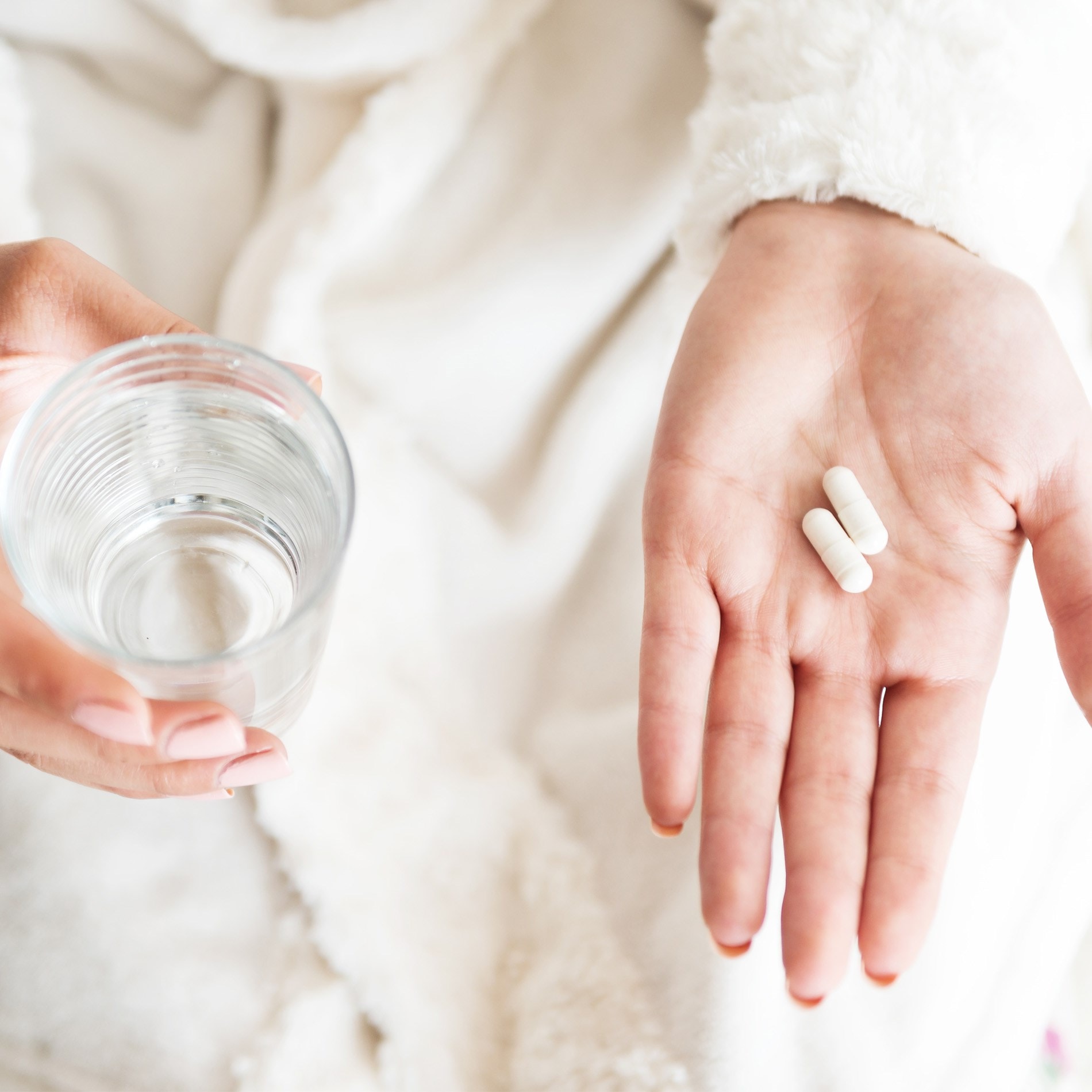 A person in a white robe holds two white capsules in one hand and a glass of water in the other.