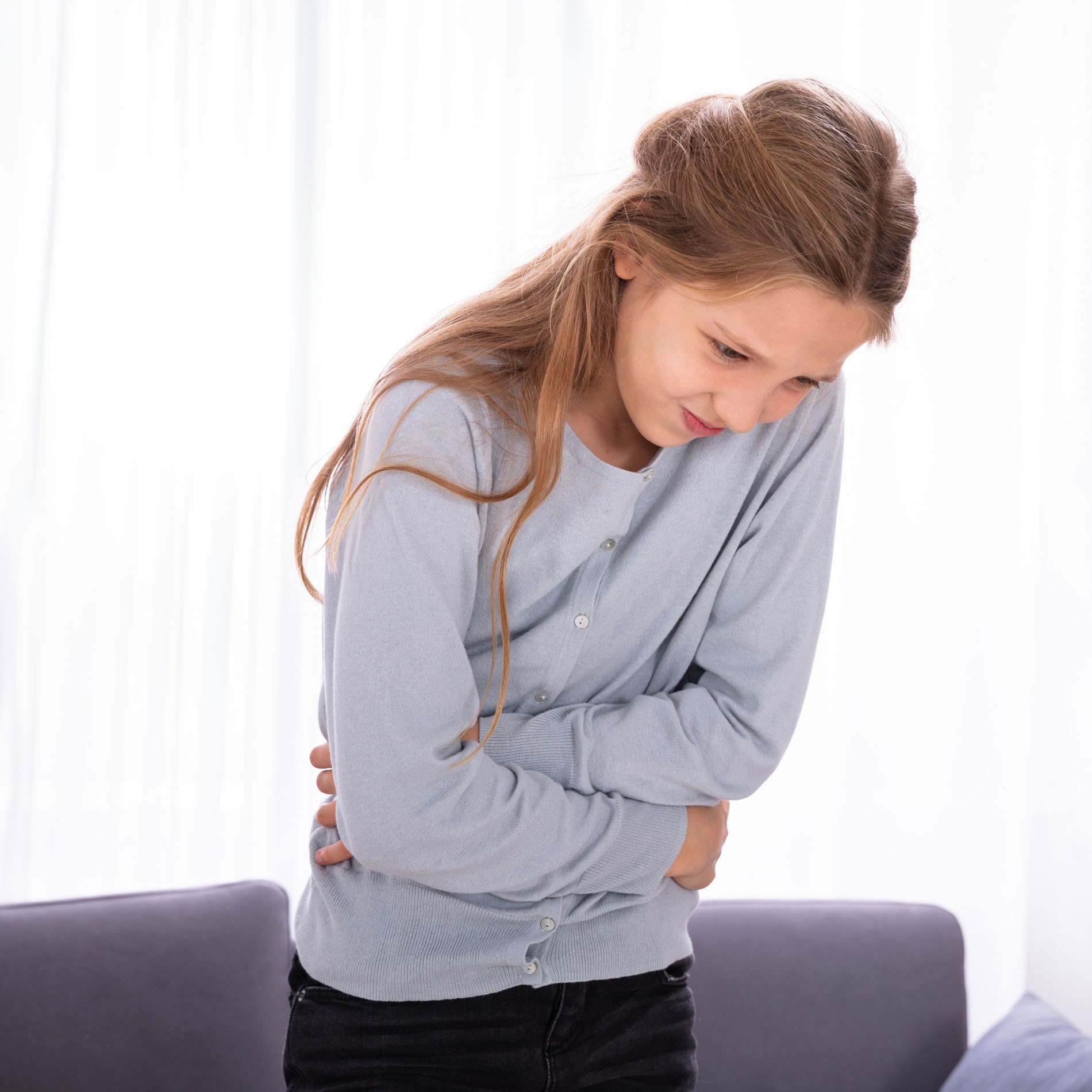 Girl in a light blue sweater clutching her stomach, appearing to be in discomfort, stands in a room with soft lighting and a sofa in the background.