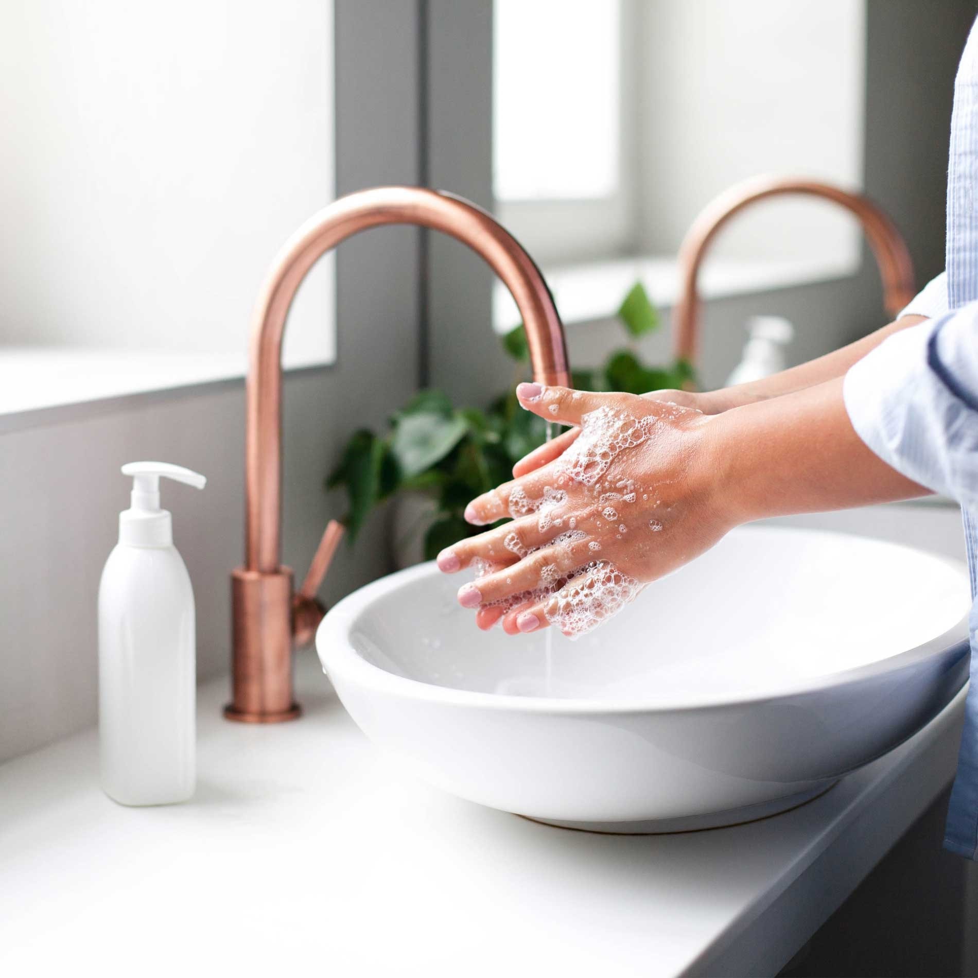 Person washing hands with soap at a white sink with a copper faucet. A soap dispenser and green plant are on the counter.