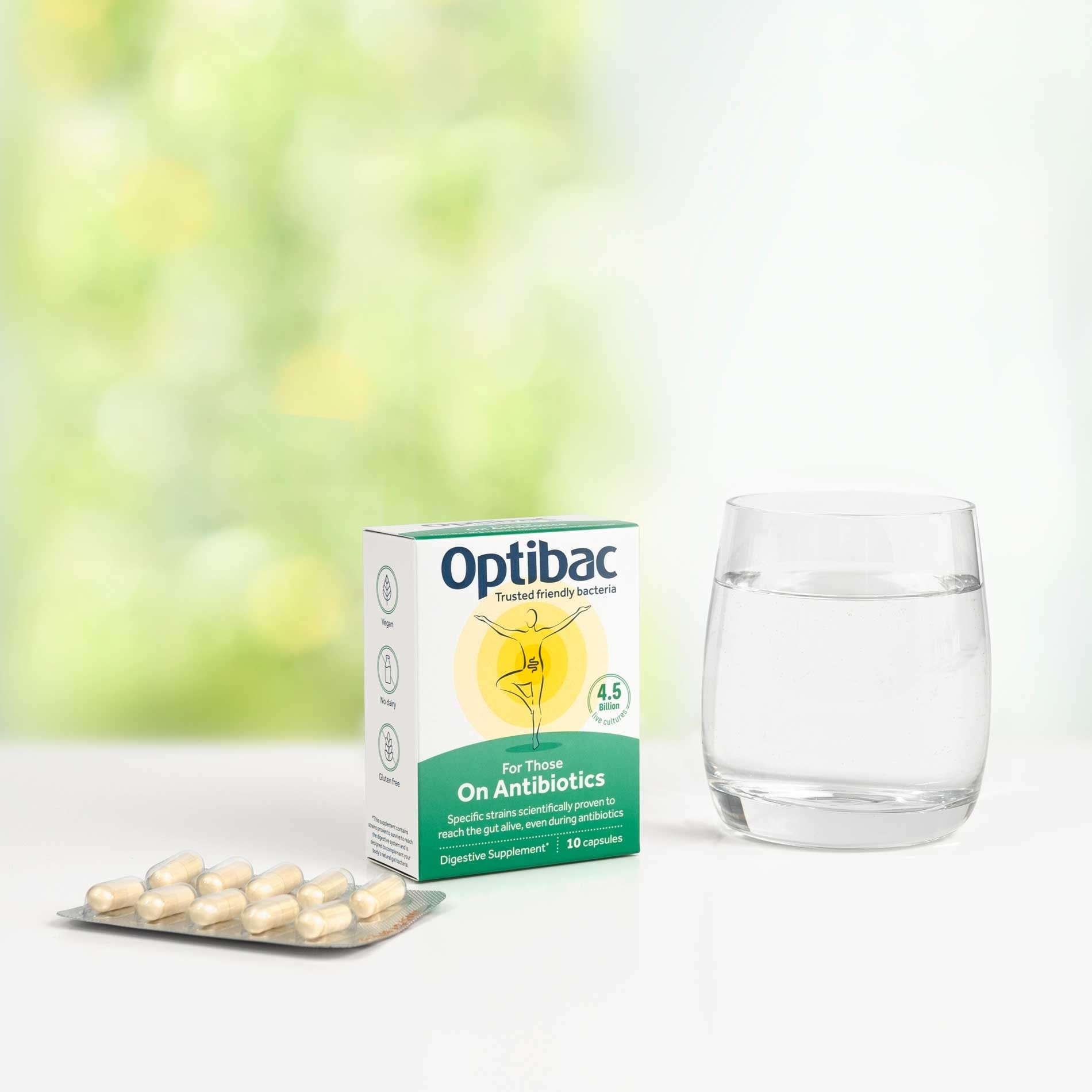 Box of Optibac probiotics for antibiotics, a blister pack of capsules, and a glass of water on a white surface with a blurred background.