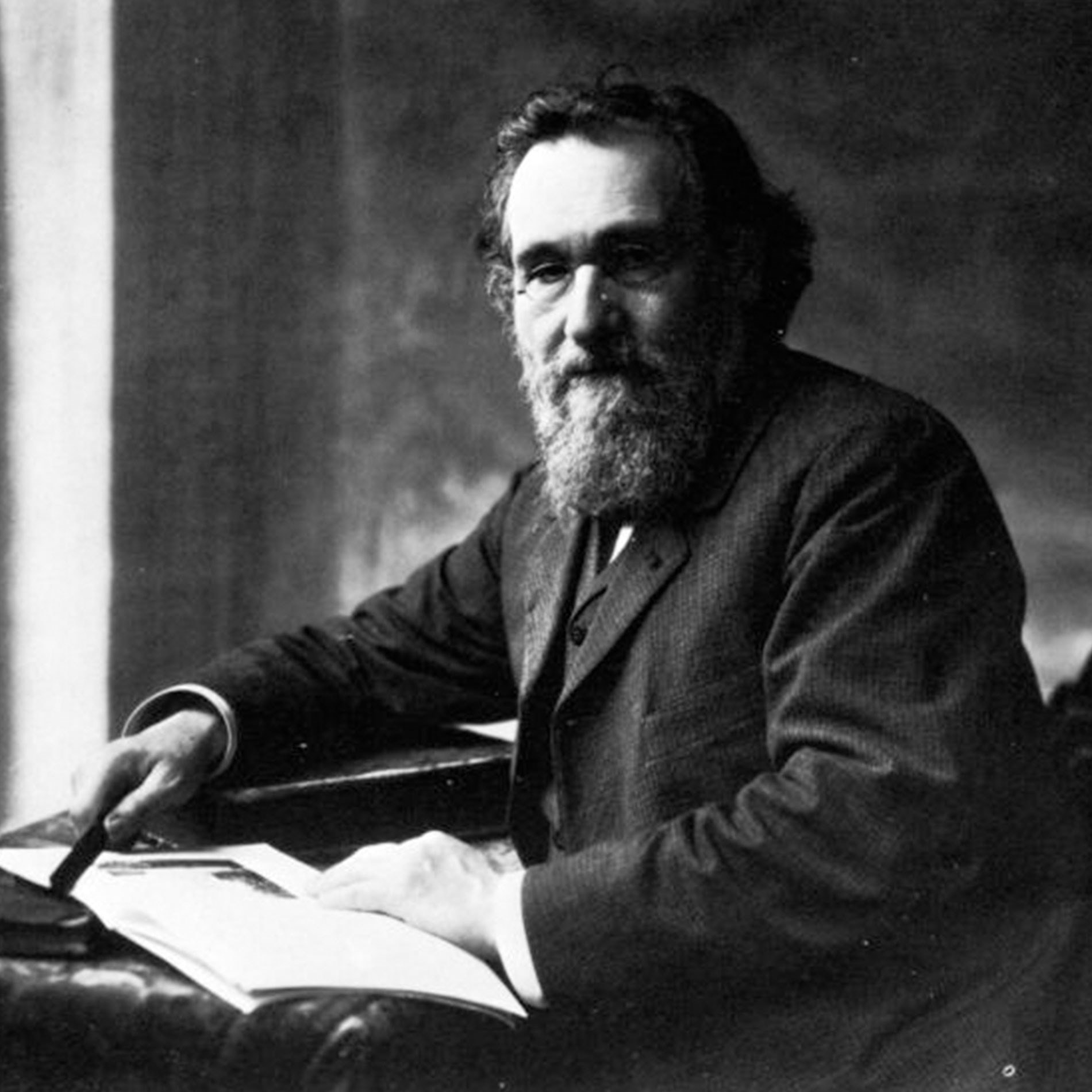 Man with a beard and glasses sitting at a desk, holding a pen, with papers in front of him. Black and white photograph.