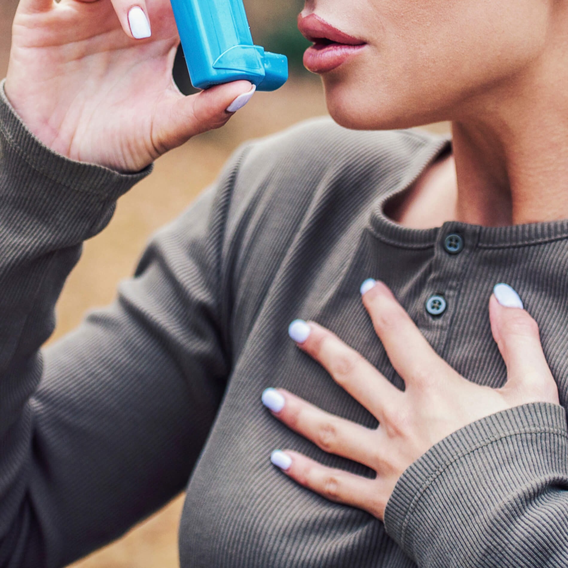 Person using a blue inhaler, wearing a gray long-sleeve shirt, with one hand on their chest.