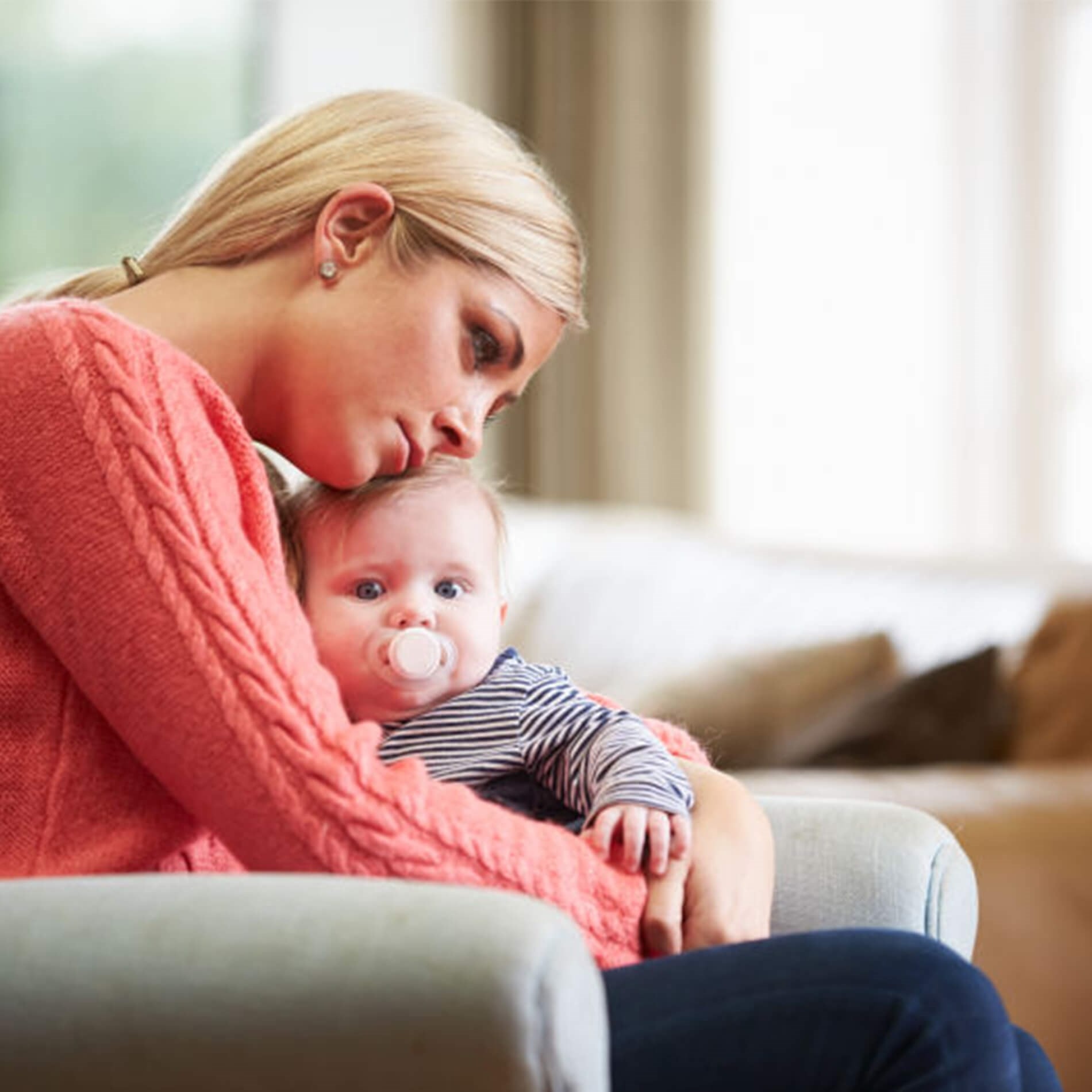 A woman in a pink sweater sits on a couch, holding a baby with a pacifier. They both look contemplative, gazing into the distance.