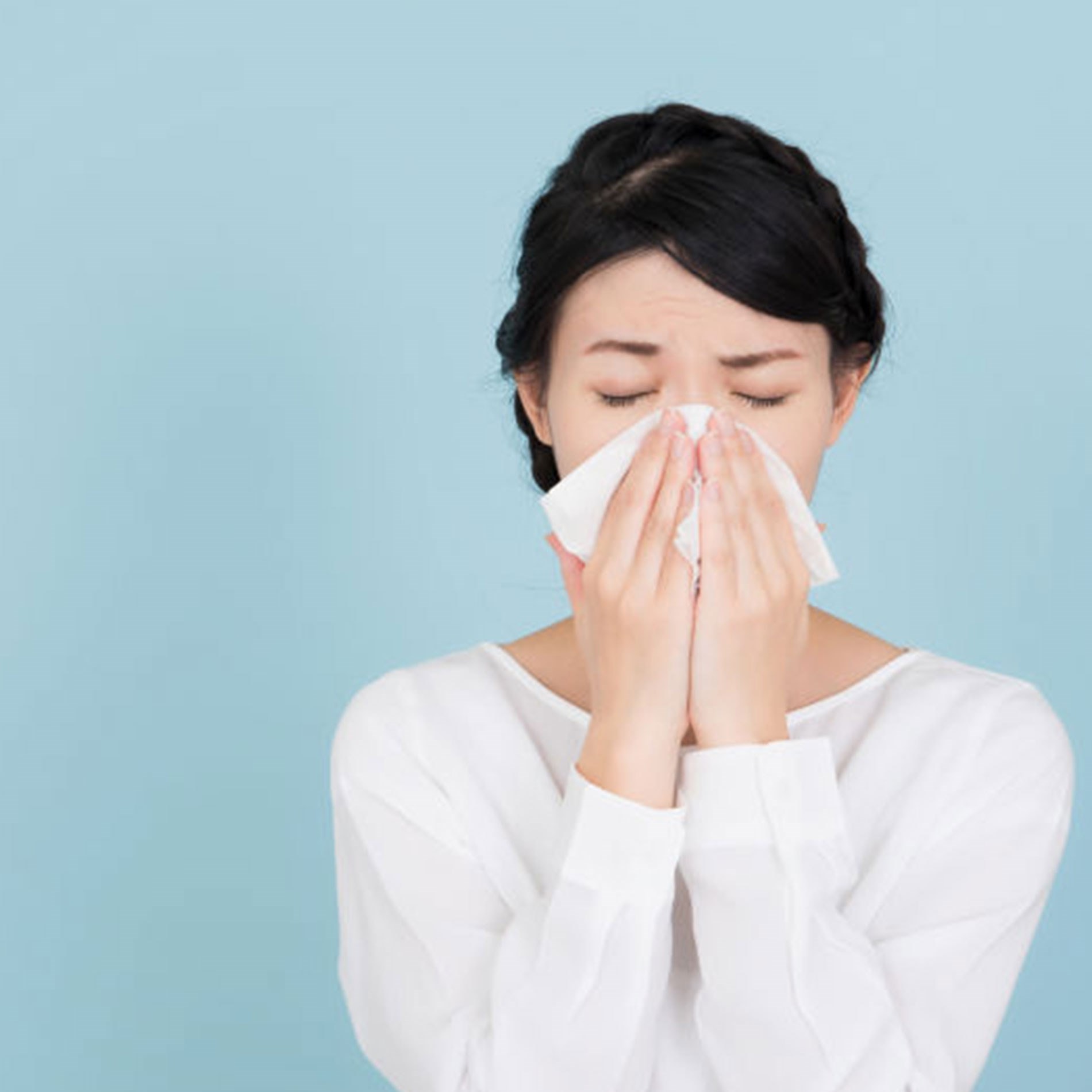 Person in a white shirt blowing their nose into a tissue against a light blue background.