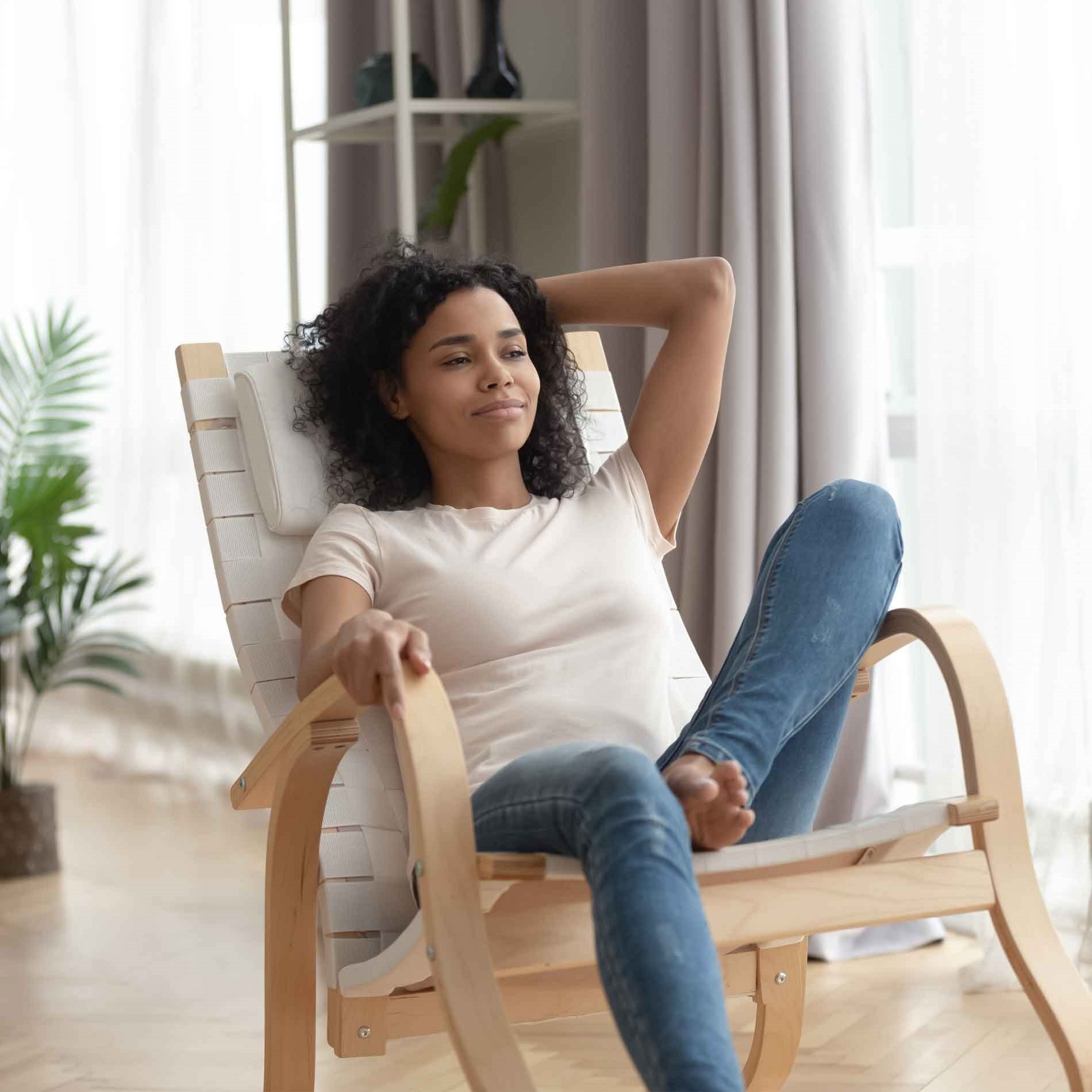 Woman relaxing in a rocking chair, wearing a white t-shirt and jeans, in a bright room with large windows and plants.