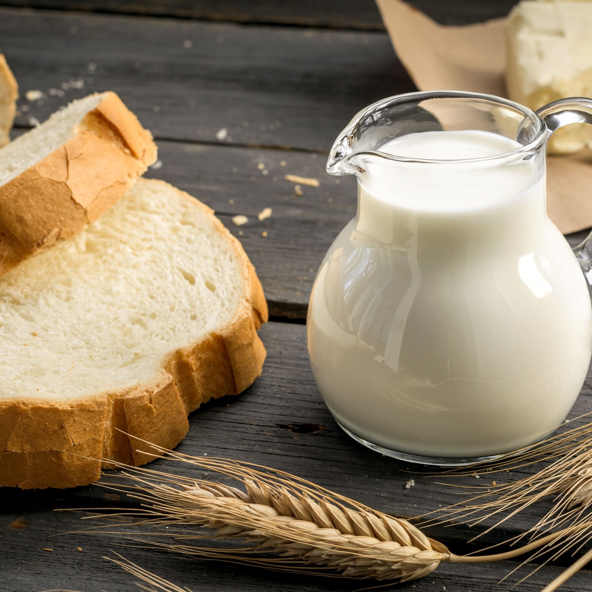 A glass pitcher of milk next to slices of bread and wheat stalks on a rustic wooden table.