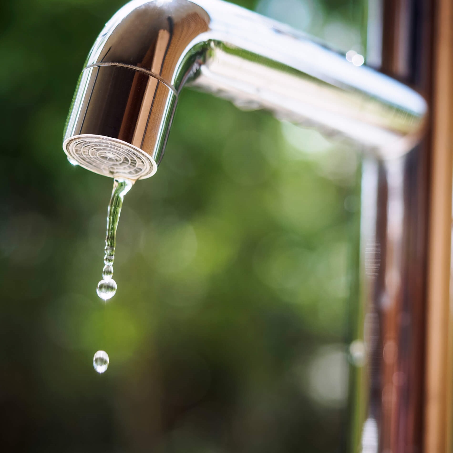 Close-up of a chrome faucet with a single water droplet falling, set against a blurred green background.