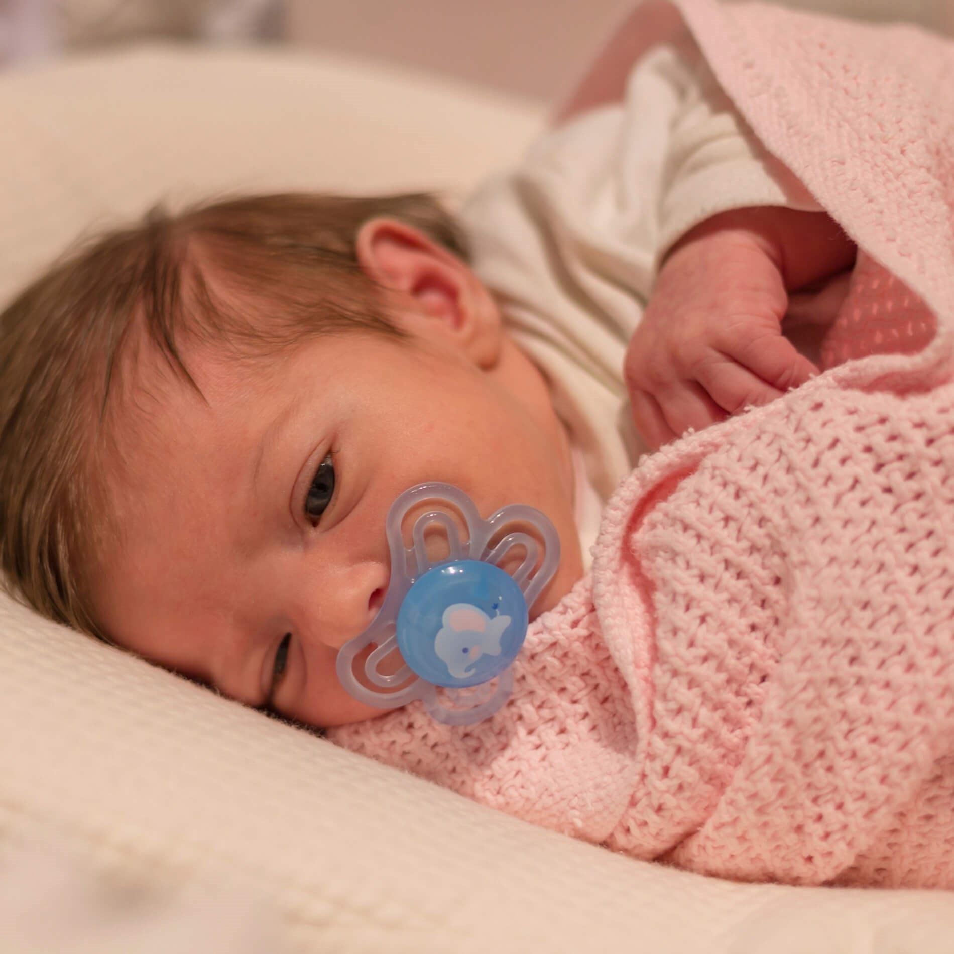 Baby lying on a soft surface, wrapped in a pink blanket, with a blue pacifier in their mouth, gazing softly.