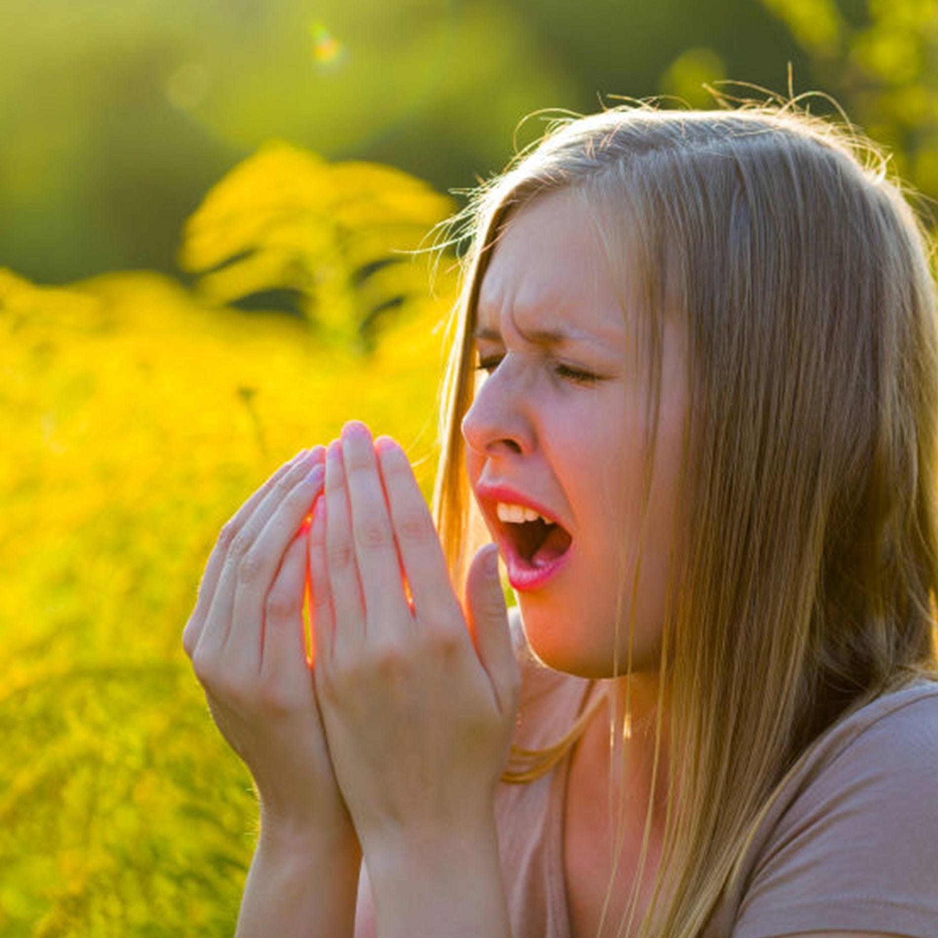 A woman sneezing into her hands in a sunny field of yellow flowers, possibly experiencing allergies.