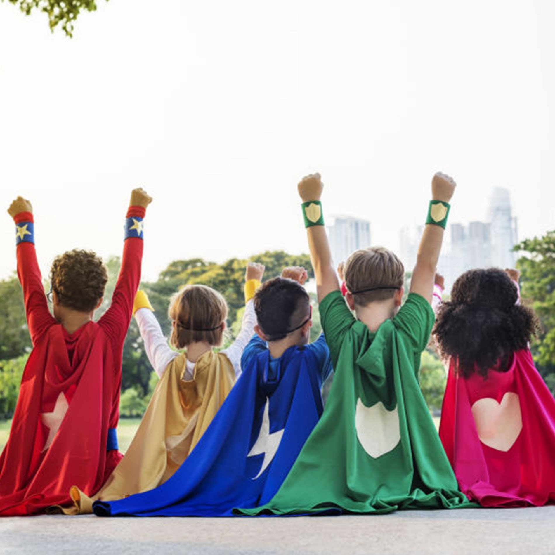 Five children in colorful superhero capes sit on the ground, raising their fists triumphantly, with a cityscape in the background.