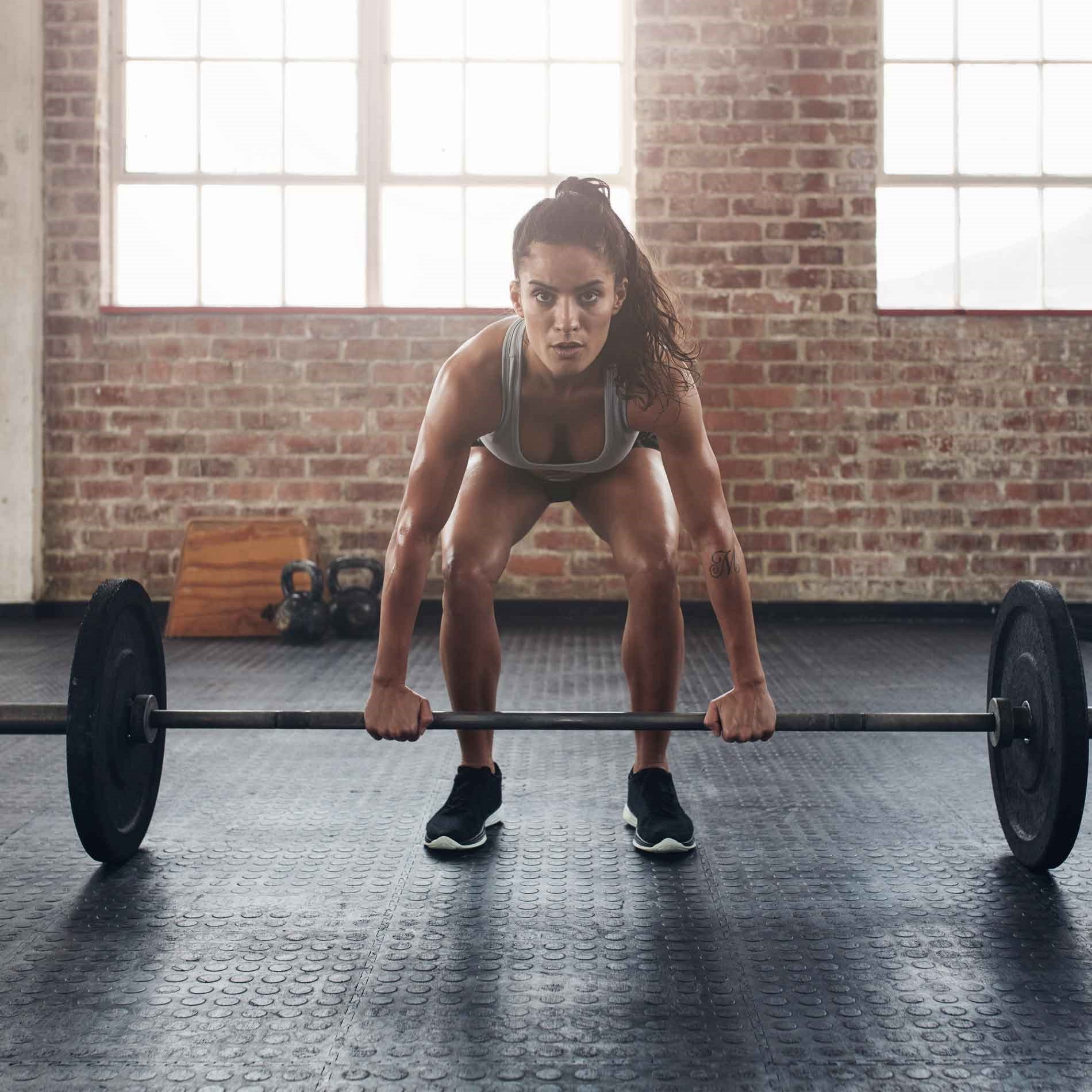 A woman in athletic wear prepares to lift a barbell in an industrial-style gym with brick walls and large windows.