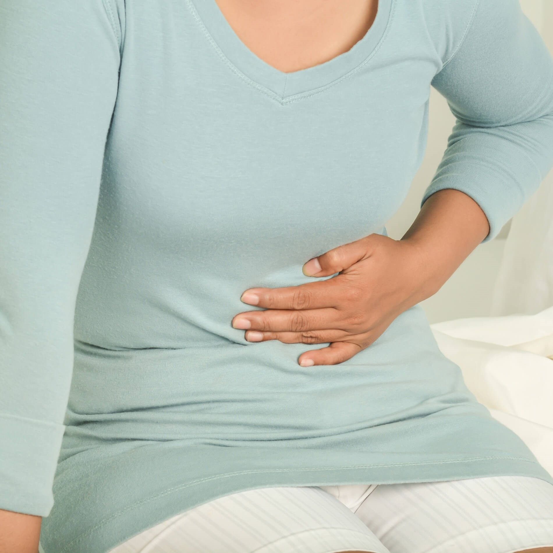 Person in a light blue shirt sitting on a bed, holding their stomach with a pained expression.