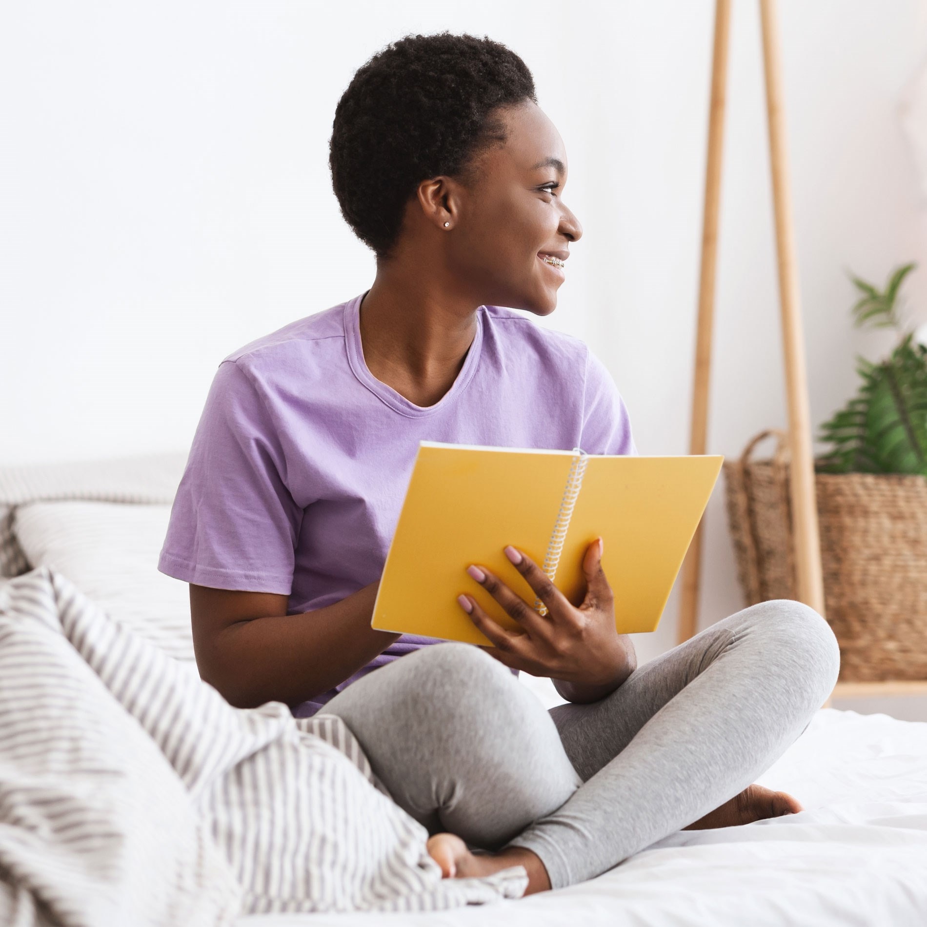 Person in a purple shirt and gray pants, sitting on a bed, smiling while holding a yellow notebook. Cozy room with plants in the background.