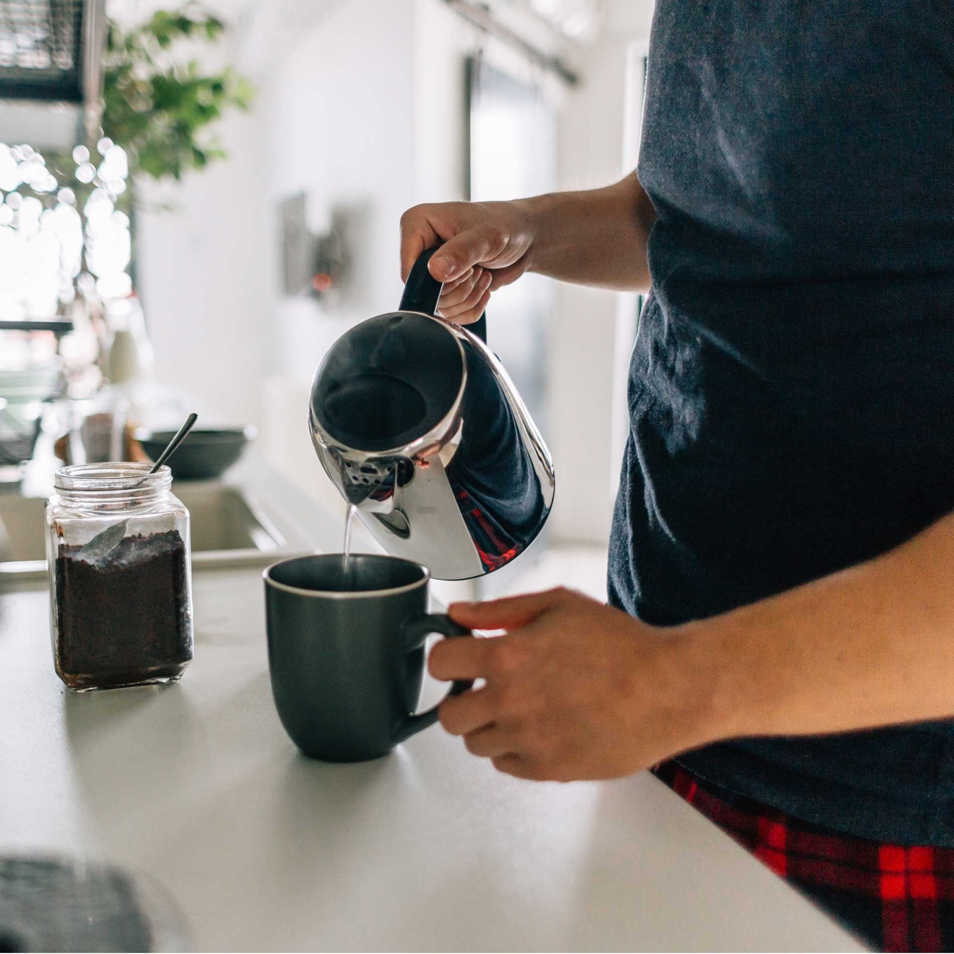 Person pouring hot water from a kettle into a mug on a kitchen counter, with a jar of coffee beside them.