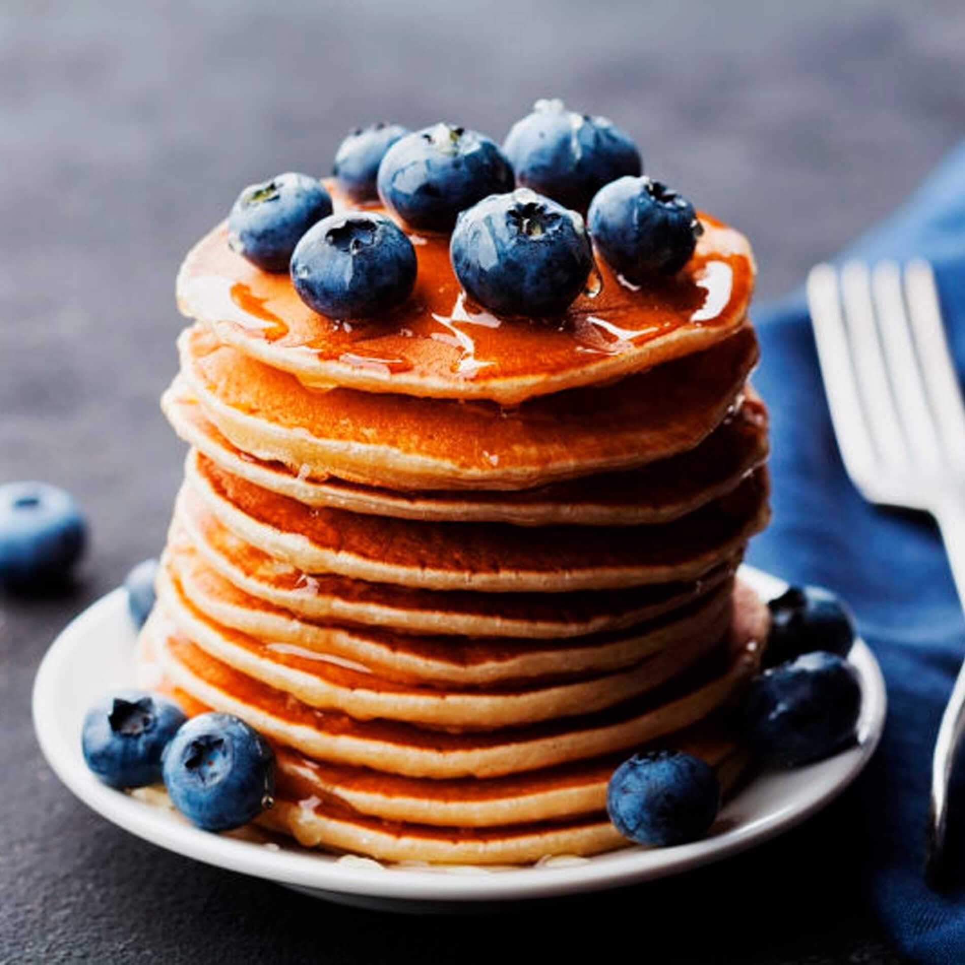 A stack of pancakes topped with fresh blueberries and syrup on a white plate, with a fork and blue napkin beside it.