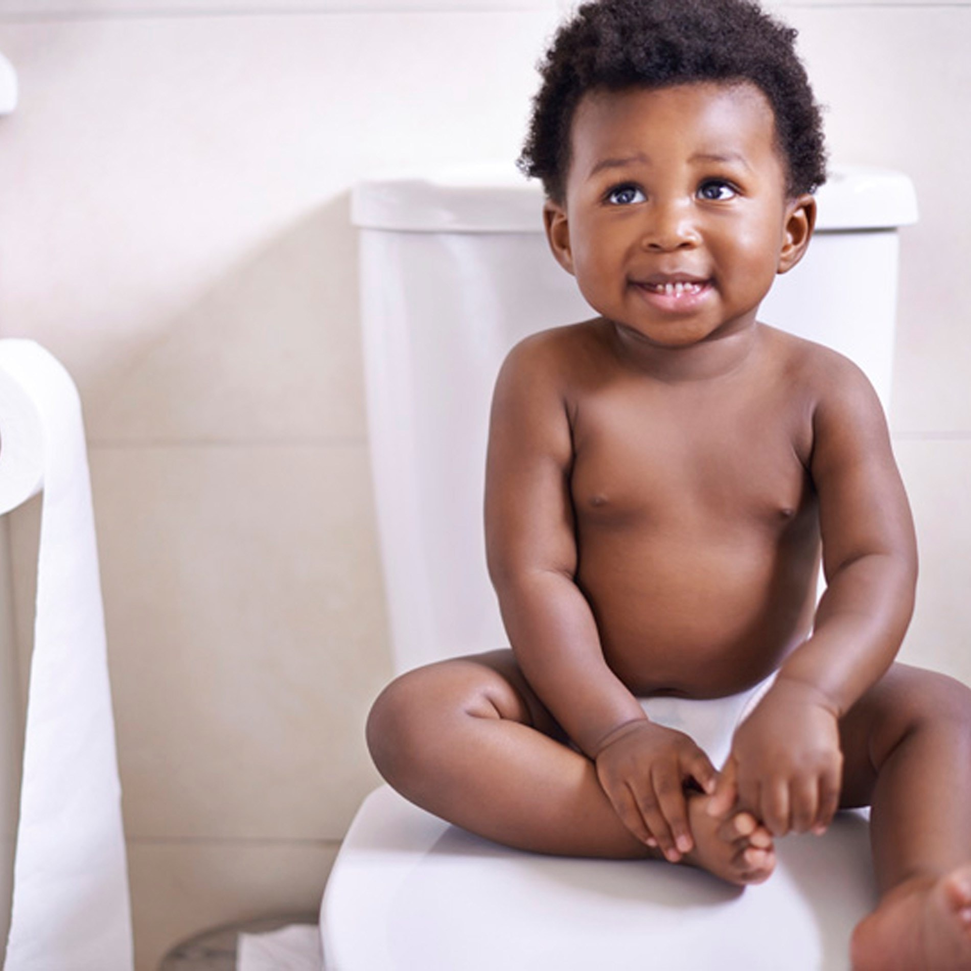 A baby with curly hair sits on a toilet seat, smiling and wearing a diaper. Toilet paper is visible in the background.
