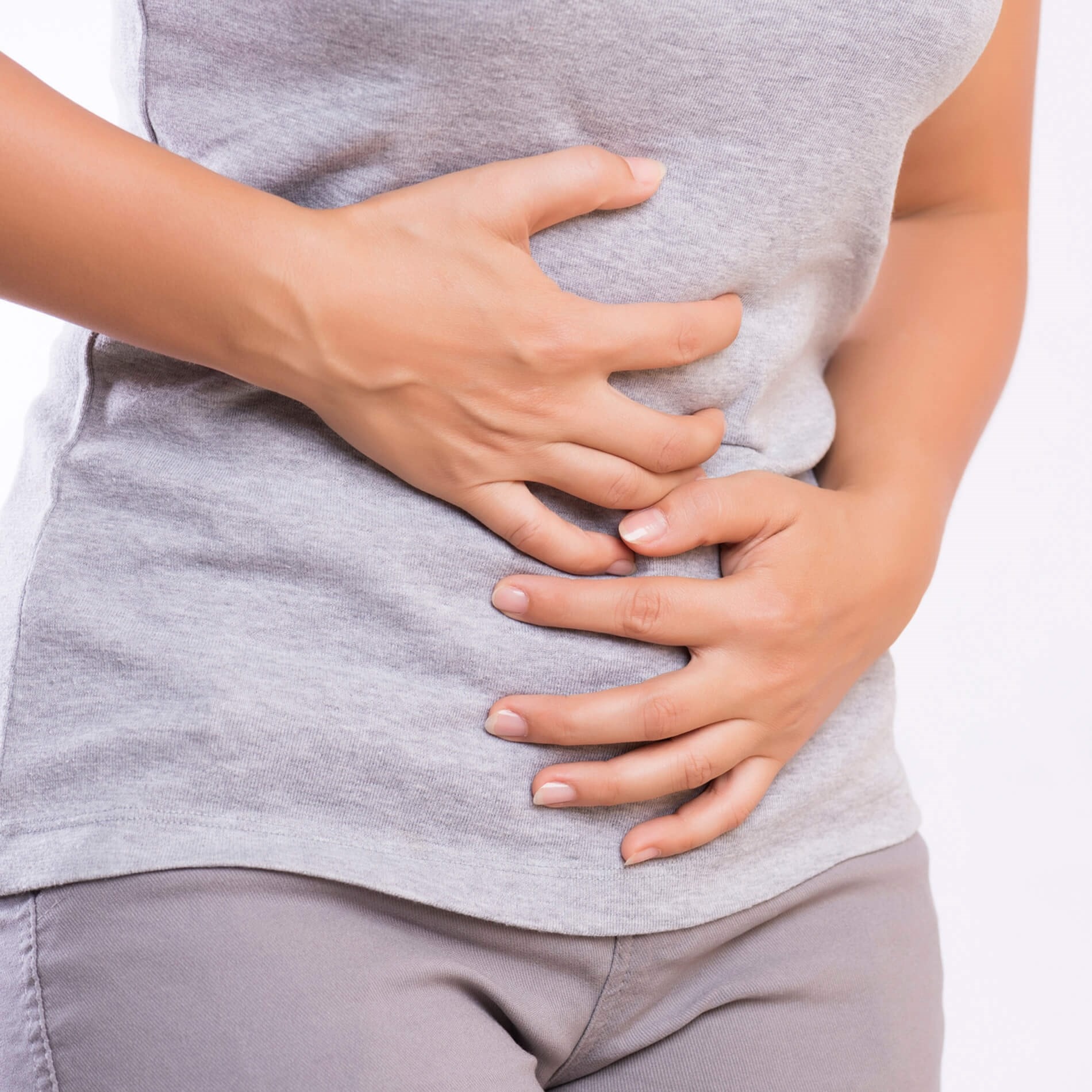 Person clutching their stomach with both hands, wearing a gray shirt and beige pants, possibly indicating discomfort or pain.