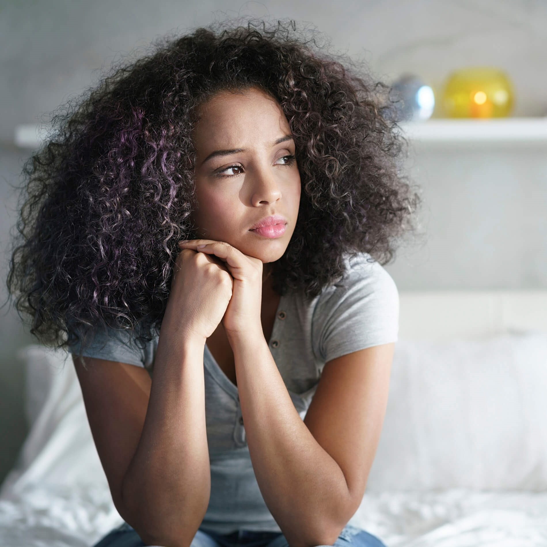 Woman with curly hair sitting on a bed, looking thoughtfully to the side, with a shelf and colorful vases in the background.
