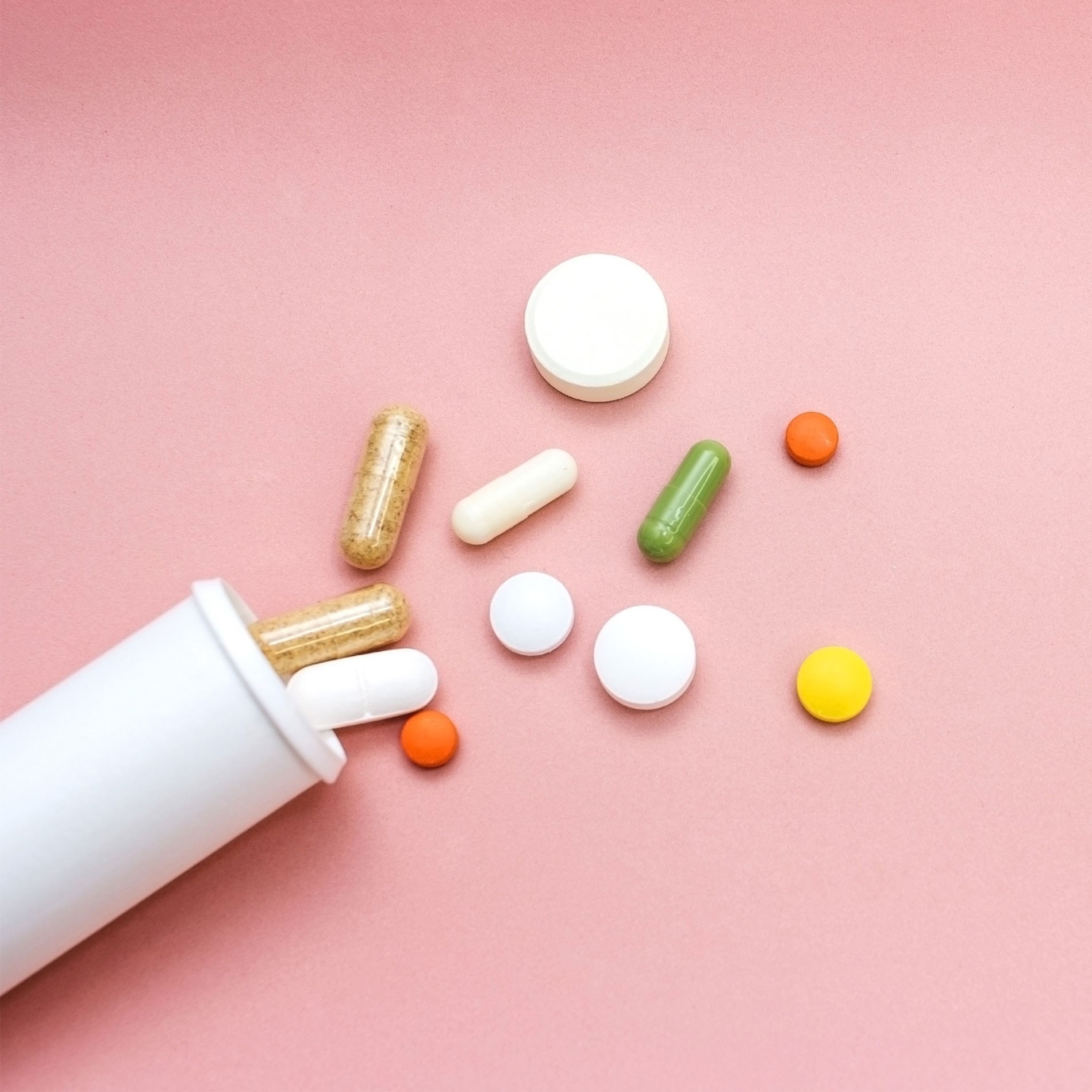 Various pills and capsules spill from a white container onto a pink surface.