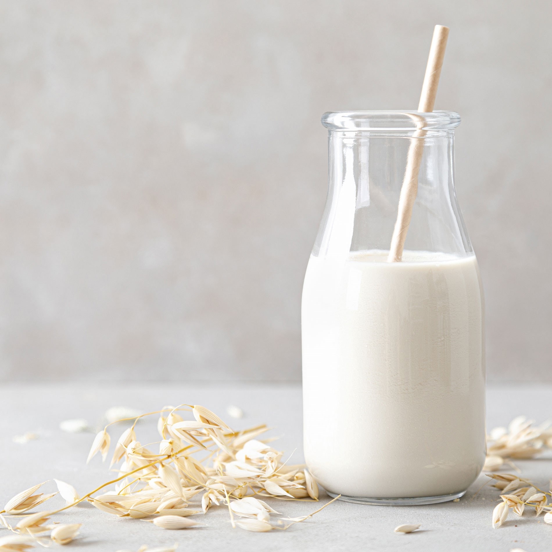 A glass bottle of oat milk with a straw, surrounded by oat husks, set on a light gray surface against a neutral background.