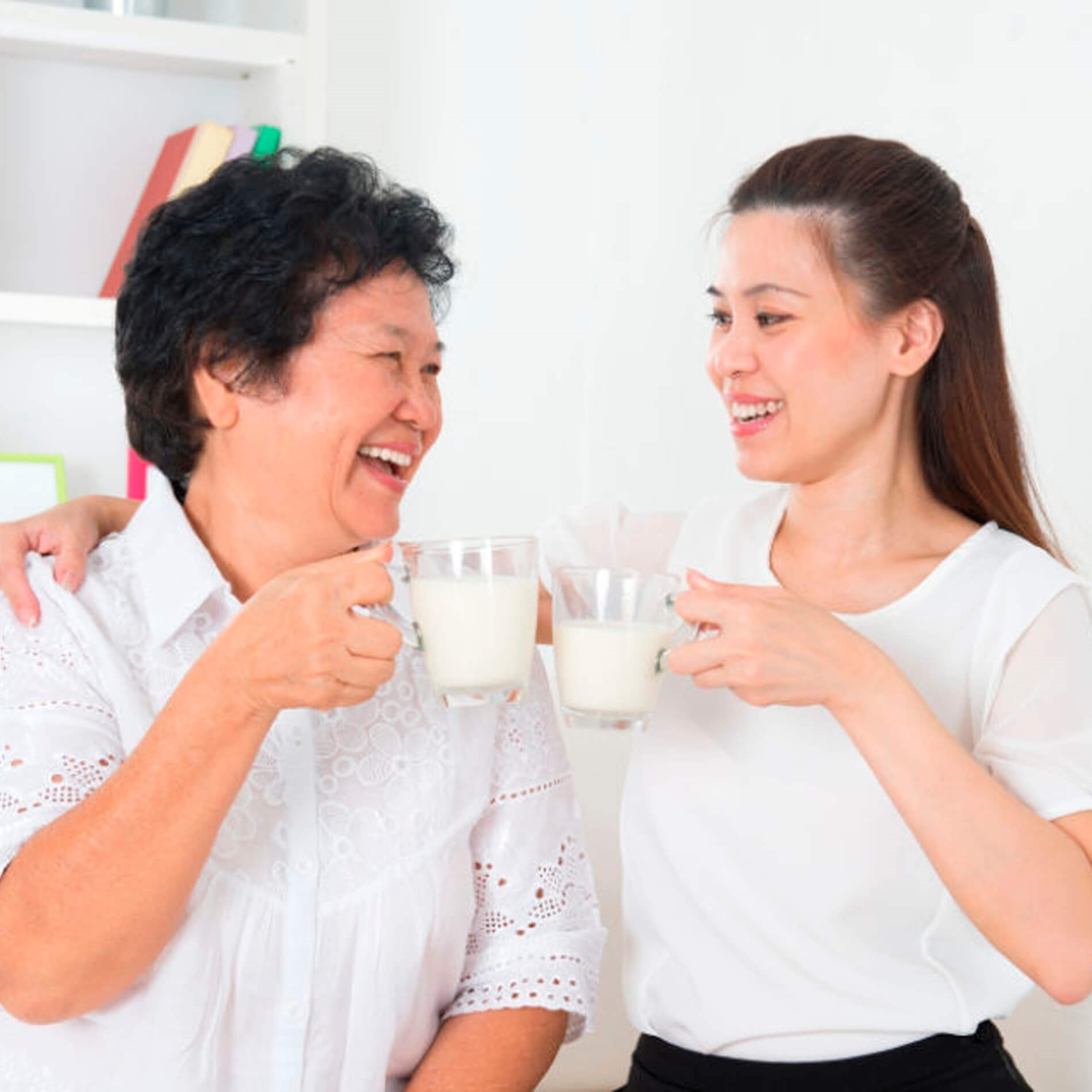 Two women smiling and clinking glasses of milk in a bright room, with books on a shelf in the background.