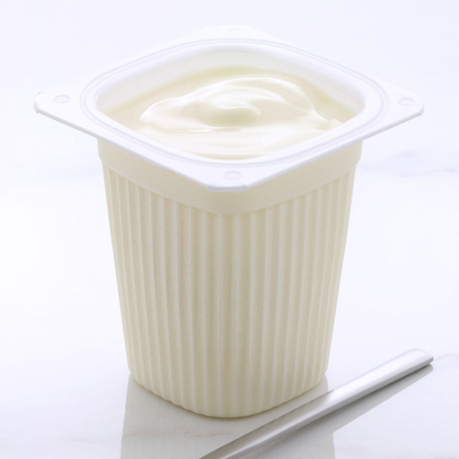 A container of creamy yogurt with a smooth surface, placed on a white background next to a silver spoon.