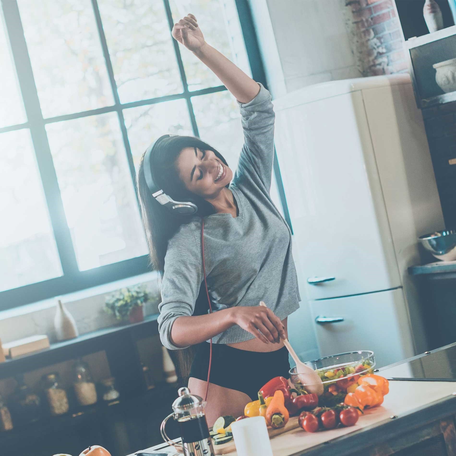 Woman wearing headphones, dancing joyfully while preparing food in a bright kitchen. Various vegetables and a French press are on the counter.
