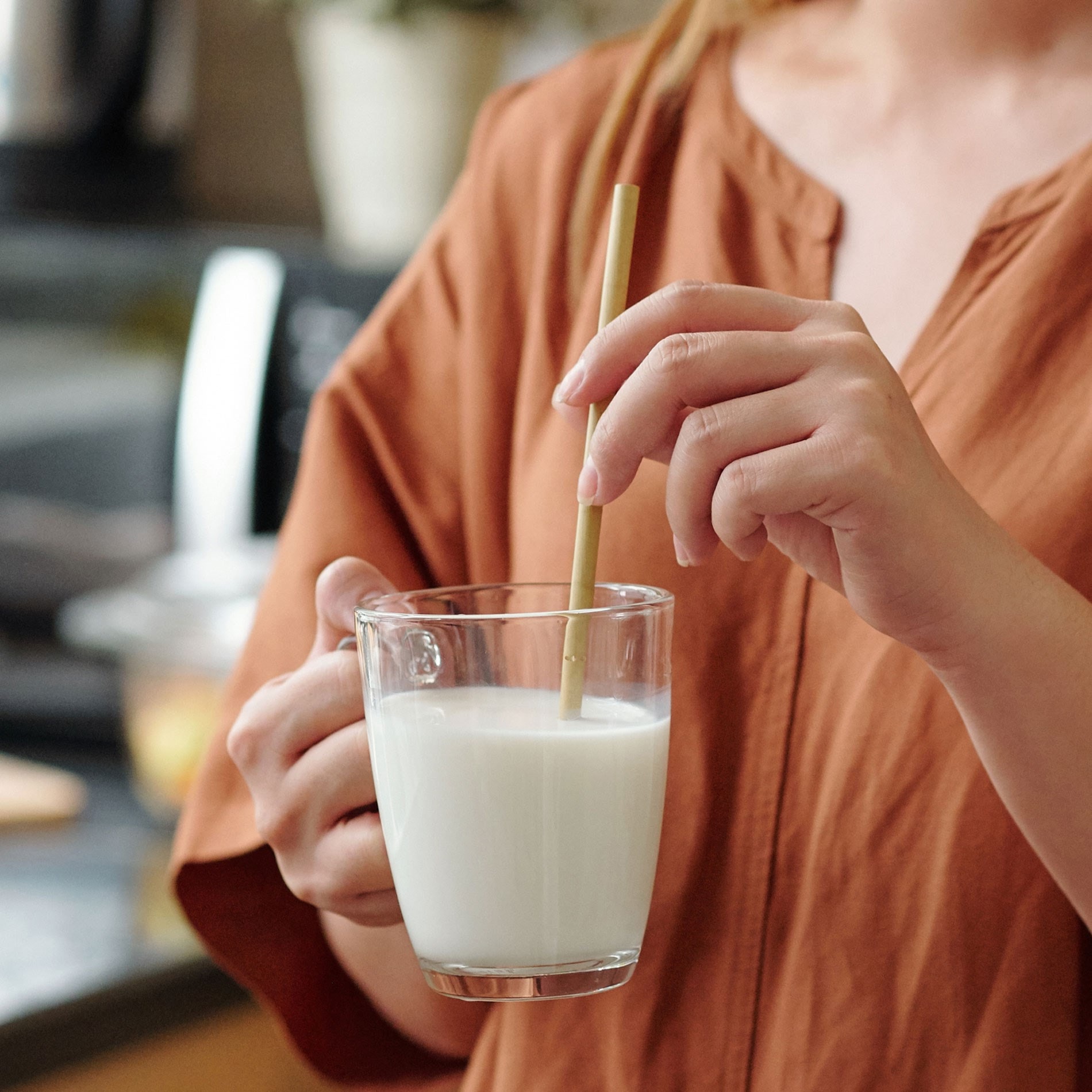 Person in a brown shirt stirring a glass of milk with a straw in a kitchen setting.