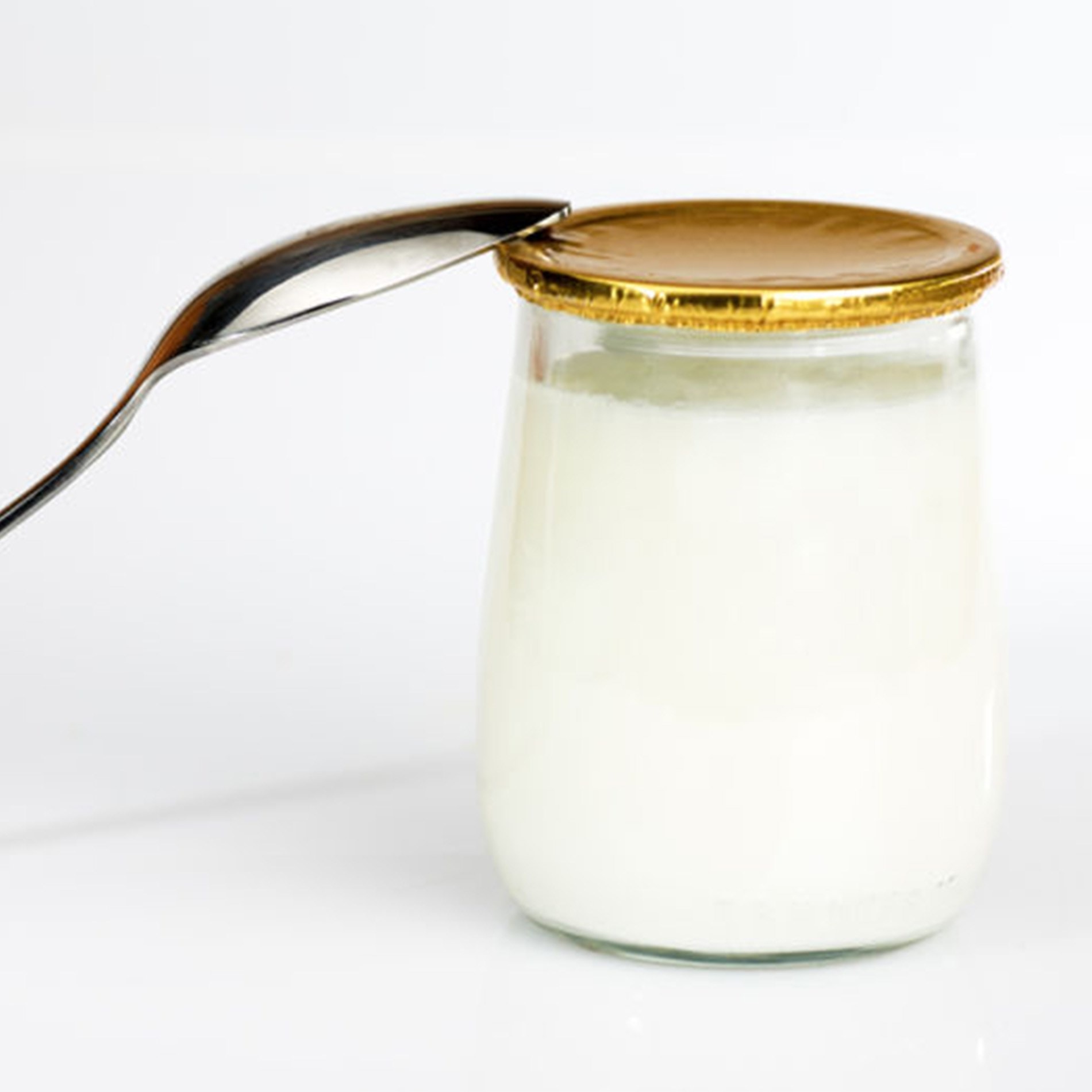 A glass jar of yogurt with a gold lid, next to a spoon, against a plain white background.