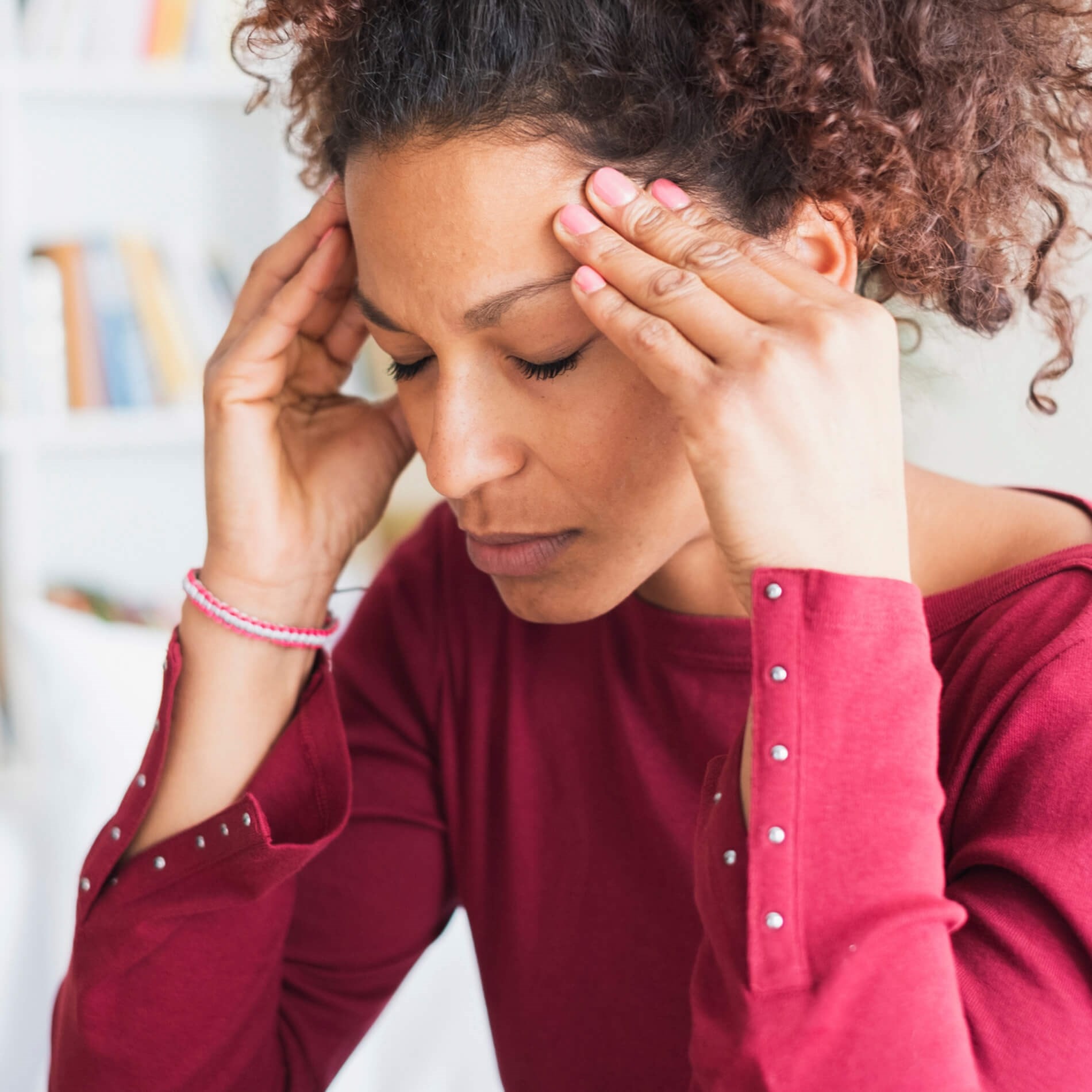 A woman in a red top holds her temples, appearing stressed or in pain, with a blurred background of bookshelves.